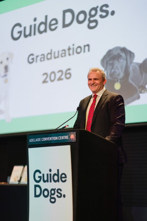 A man in a suit stands at a lectern labelled Guide Dogs at the Adelaide Convention Centre, smiling, with a screen behind him that reads Guide Dogs. Graduation 2026 and displays two loyal guide dogs.