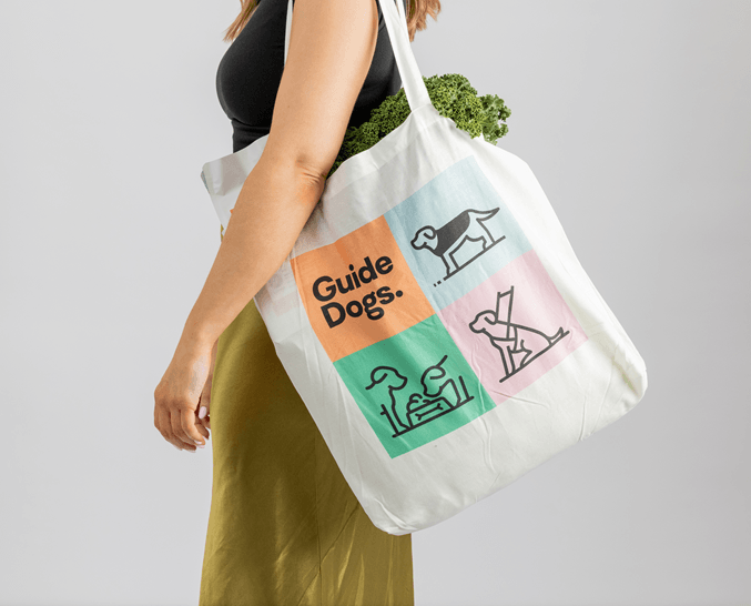 A woman carries a white tote bag with colourful Guide Dogs illustrations and text, containing leafy green vegetables. She wears a black top and olive skirt against a plain background.