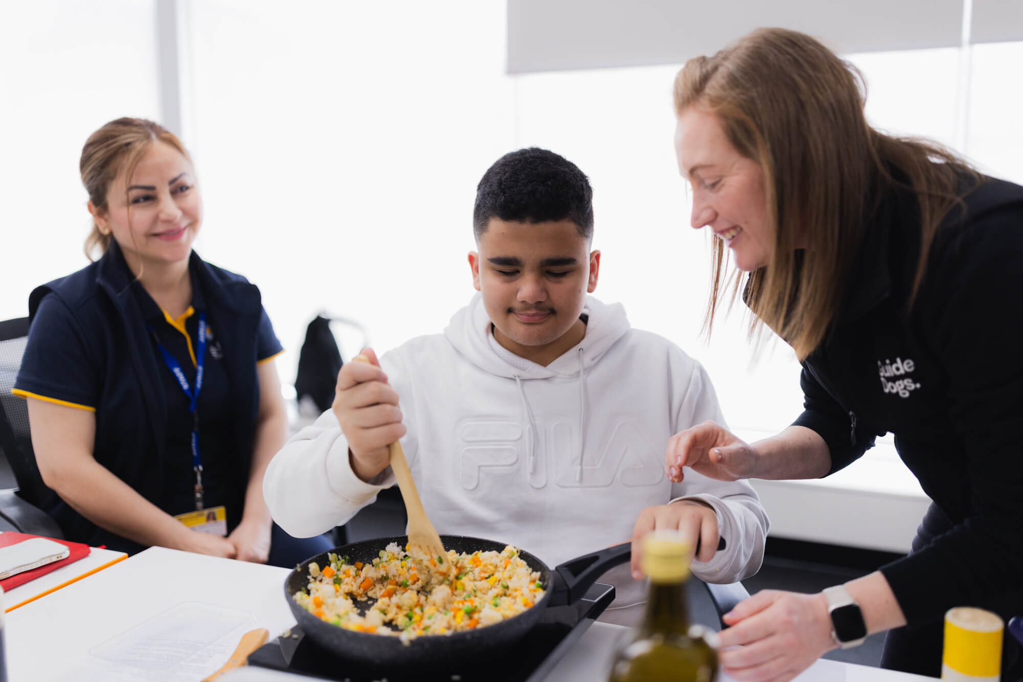 A smiling woman helps a boy in a white hoodie stir a pan of mixed vegetables, while another woman watches and smiles in the background. They appear to be taking part in a cookery activity together.