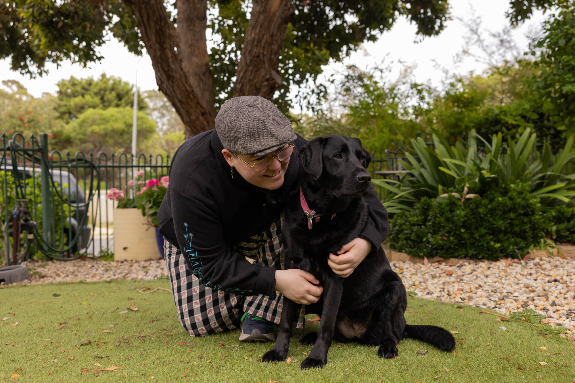 A person wearing a cap and glasses kneels on grass, smiling and hugging a black dog with a pink collar in a garden with trees and greenery.