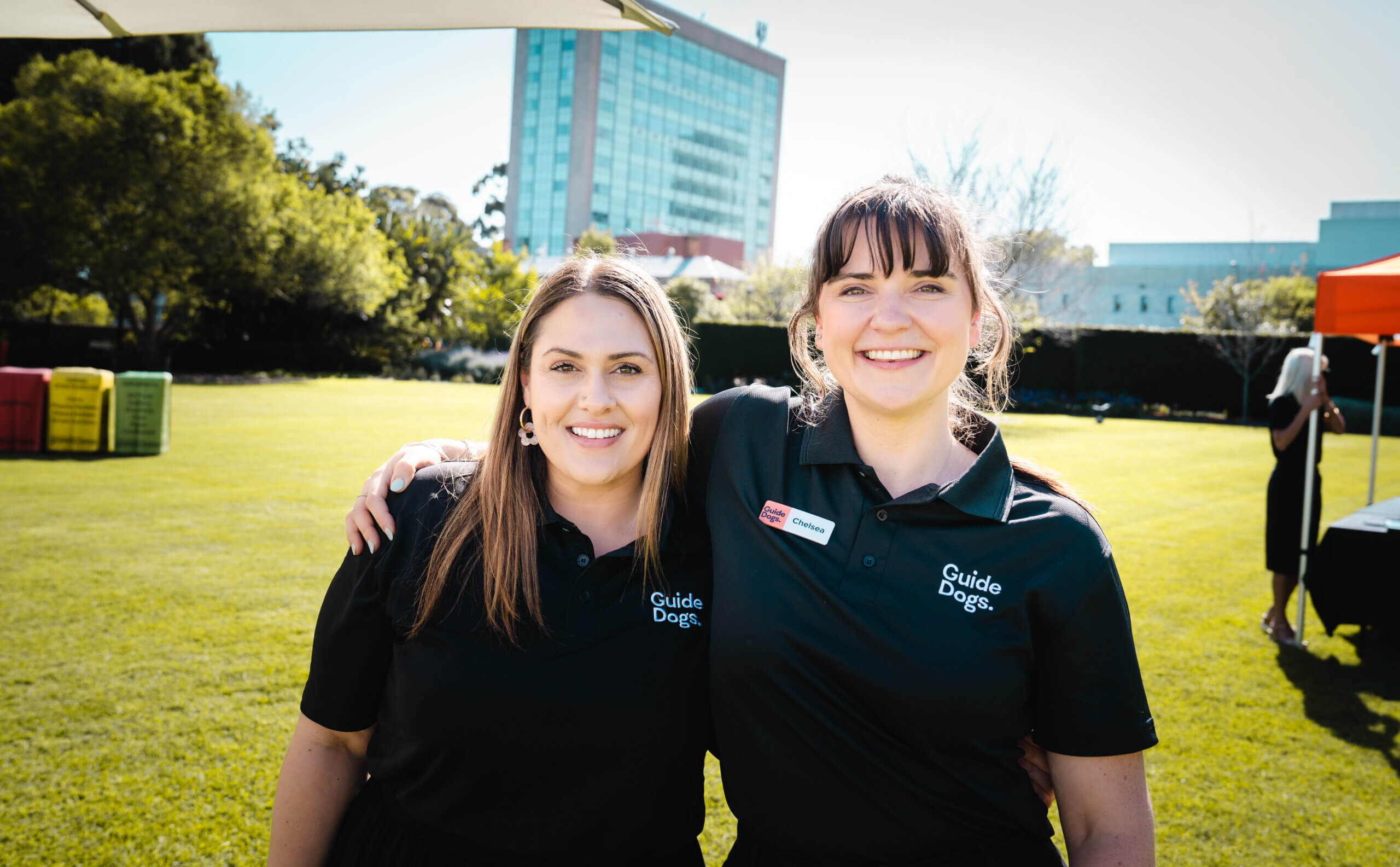 Two women wearing black Guide Dogs shirts stand on grass, smiling with arms round each other on a sunny day. A modern building, trees, and event marquees are visible in the background.