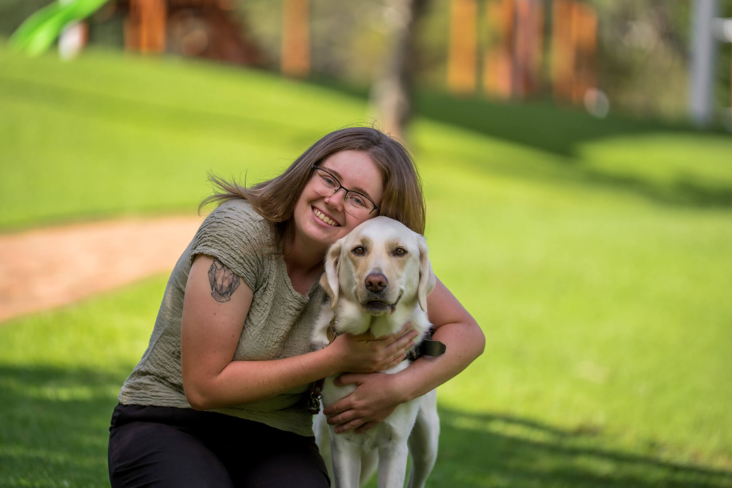 A smiling woman with glasses hugs a yellow Labrador retriever while kneeling on green grass in a sunlit park. Trees and a blurry path are visible in the background.