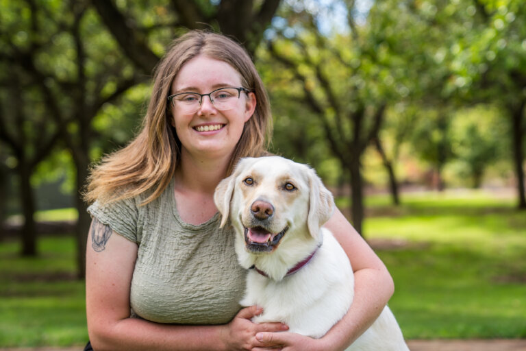A smiling woman with glasses and a light green shirt sits outdoors in a park, cuddling a happy white dog with tan ears. Trees and greenery are visible in the blurred background.