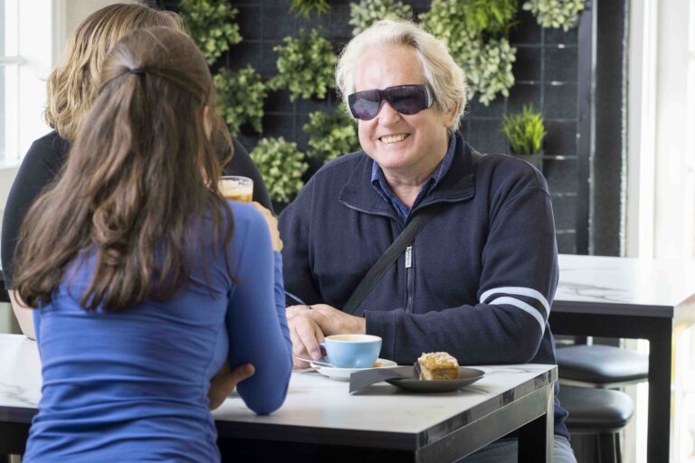 A man wearing sunglasses and a dark jacket smiles whilst chatting with two women at a café table. There are drinks, a coffee cup, and a pastry on the table. Green plants decorate the wall behind them.