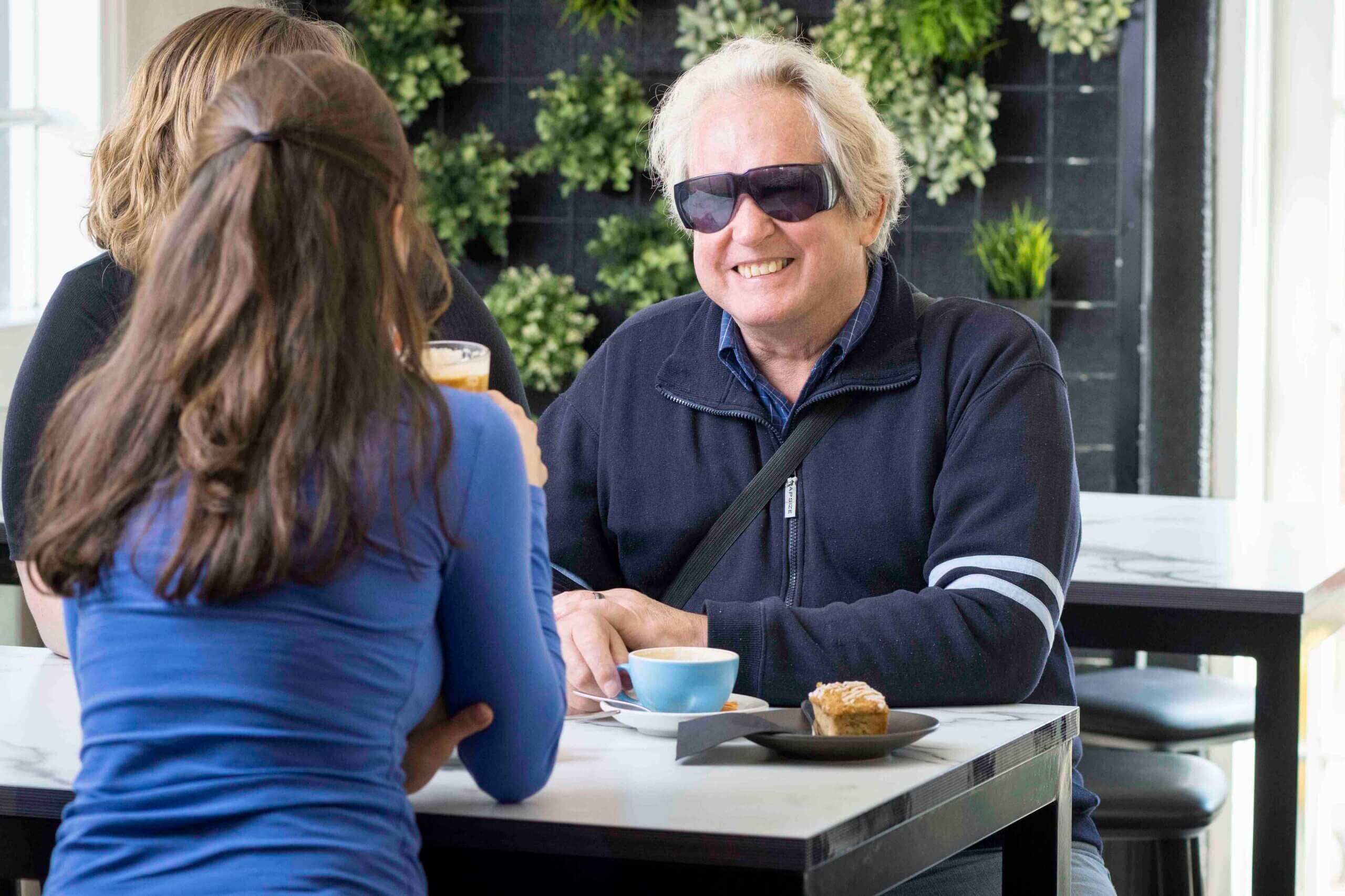 A man wearing sunglasses smiles while sitting at a café table with two women, enjoying a coffee and pastry. Green plants create a lush backdrop on the wall behind them.