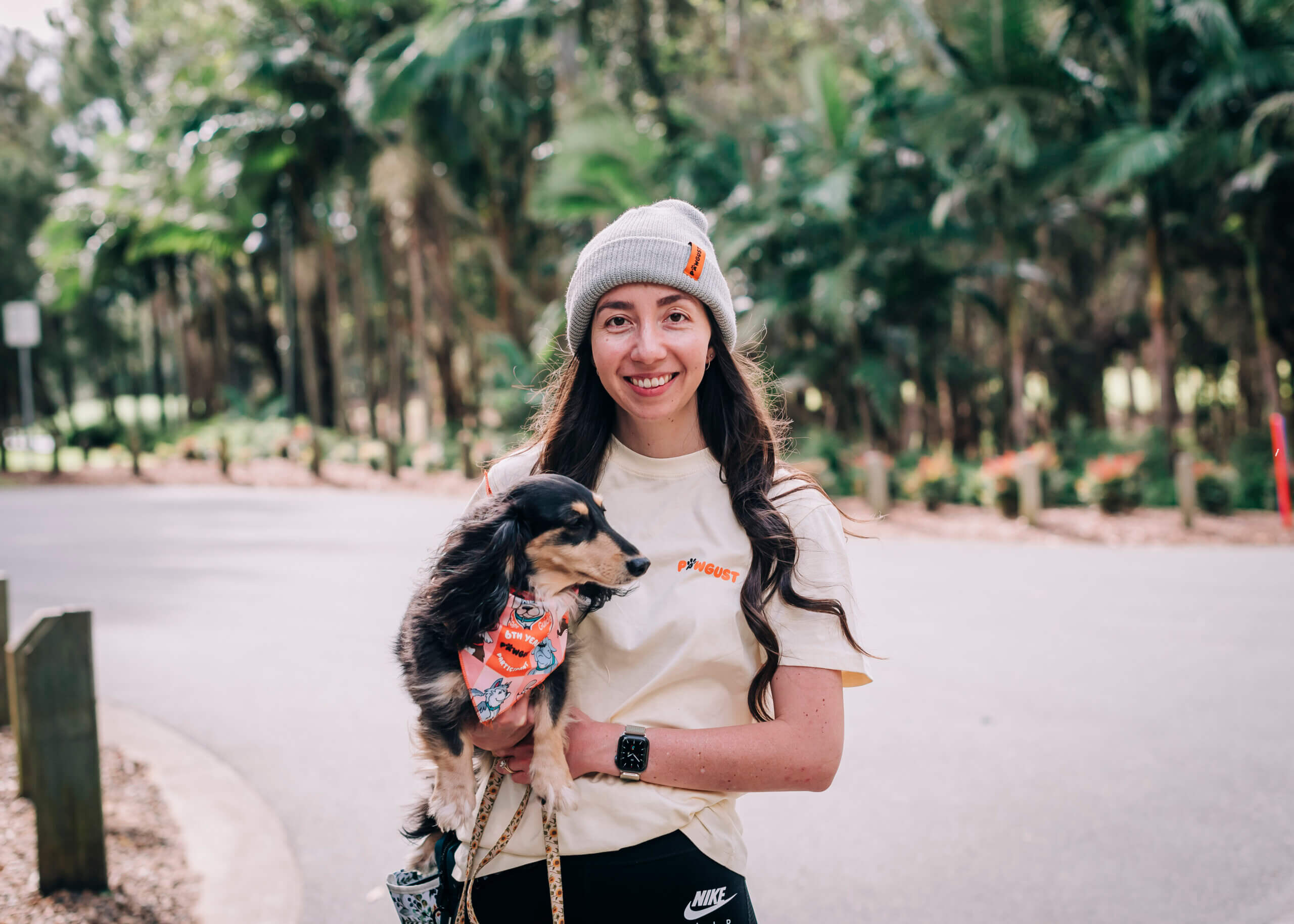 A smiling woman in a grey beanie and casual clothes holds a small dog wearing a colourful bandana on a lead, standing on a road with lush green trees in the background.
