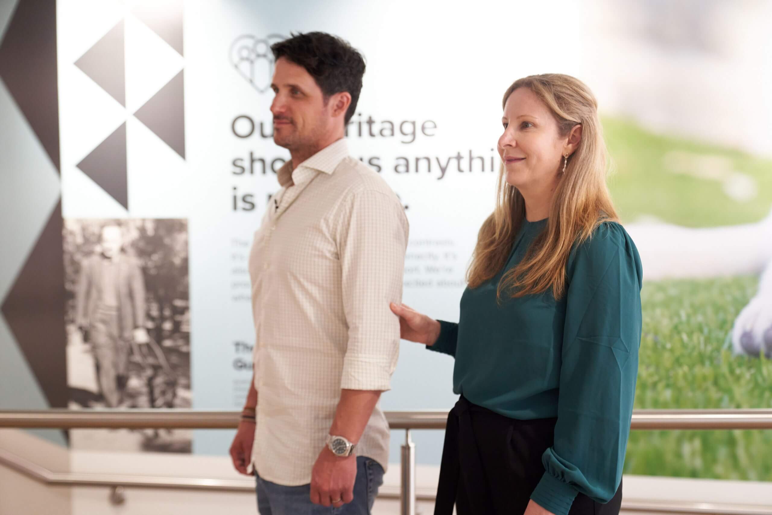 A woman with long blonde hair stands next to a man with dark hair, gently touching his back, in a brightly lit room with informational displays on the wall behind them.