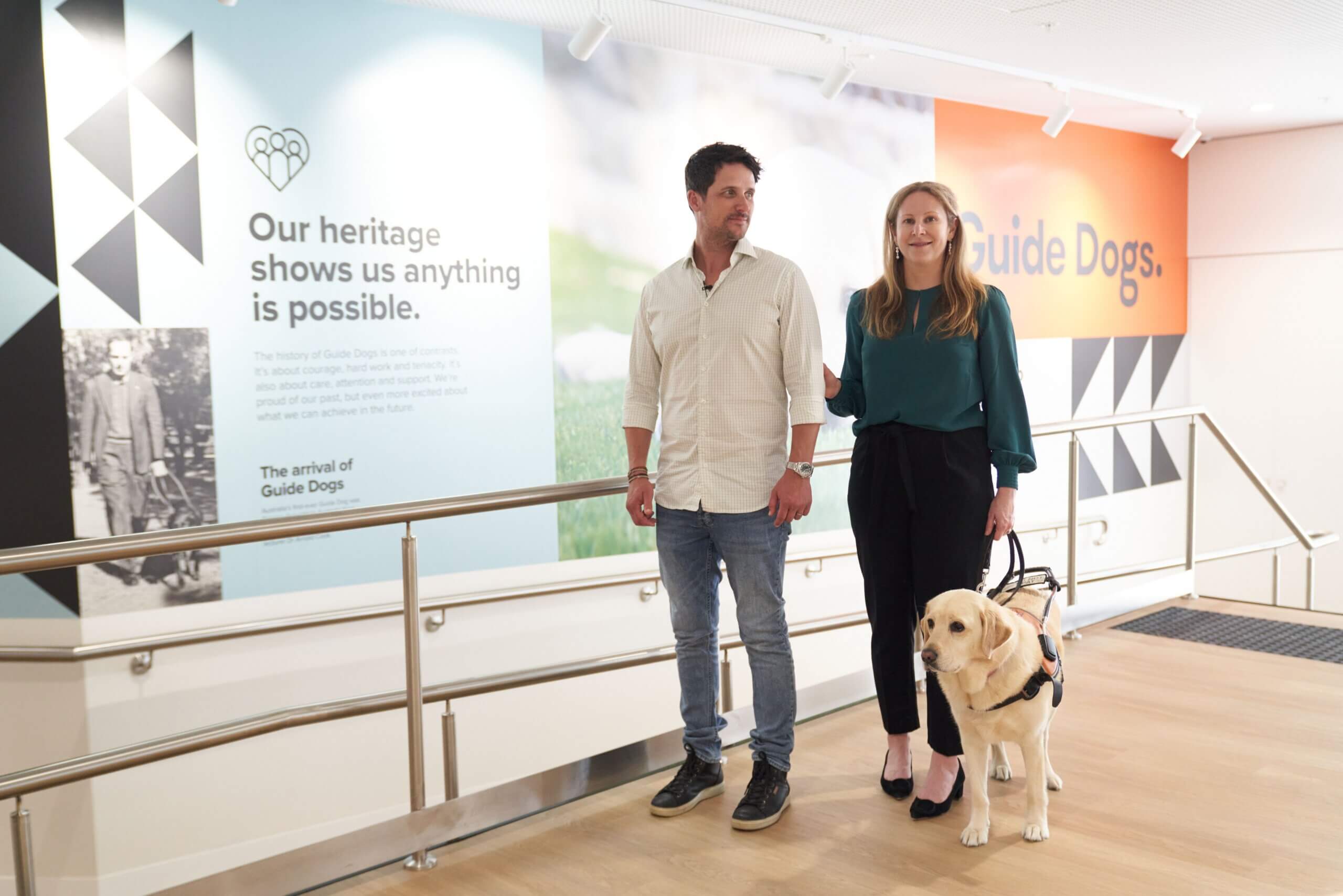 A man and woman stand side by side indoors, with the woman holding a harness attached to a guide dog. Behind them is a wall display about the history of guide dogs, featuring text and images.