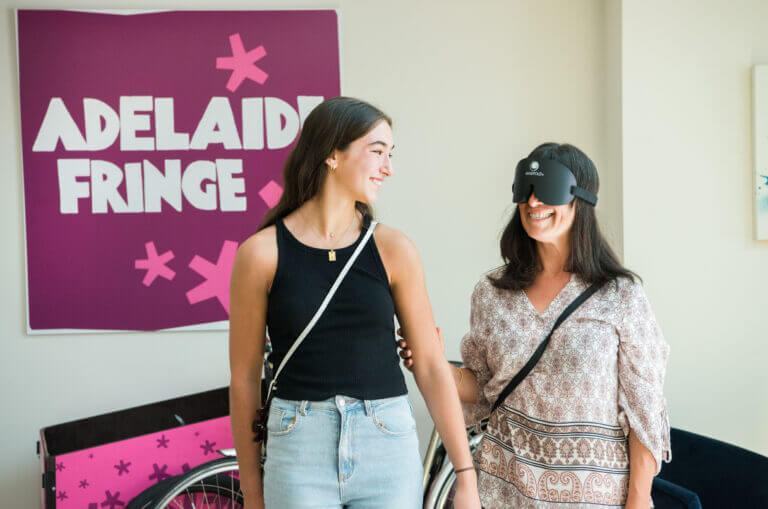 Two women smile and walk together; one wears a black blindfold. They stand in front of a purple Adelaide Fringe sign. Both have crossbody bags and casual outfits.