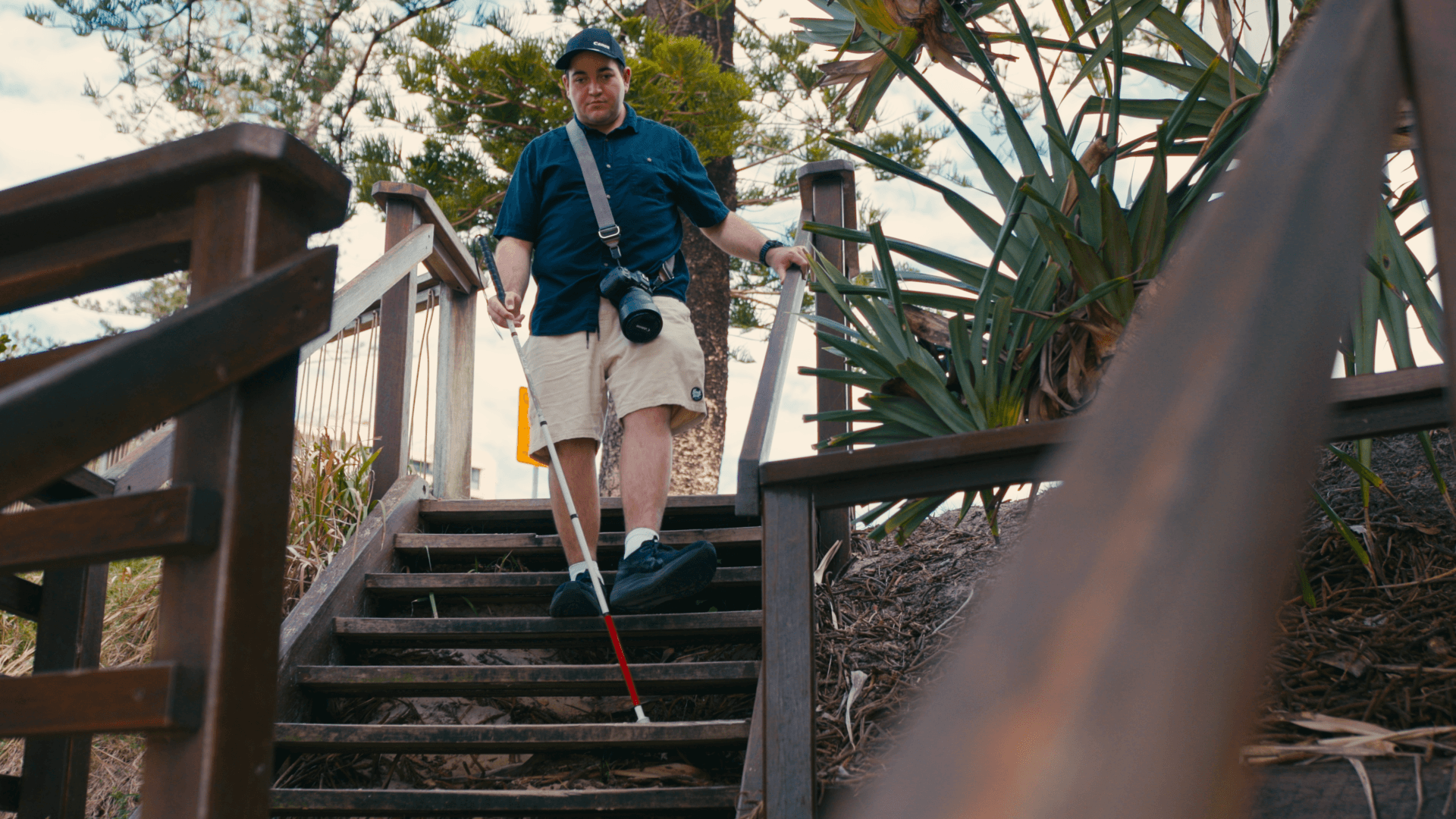 A man with a white cane walks down outdoor wooden stairs, holding the handrail for support. He wears a navy shirt, beige shorts, a cap, and a camera over his shoulder. Lush greenery surrounds the stairs.