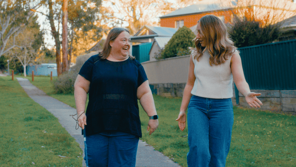 Two women are walking and talking on a suburban pavement lined with trees and houses, smiling and enjoying each other's company on a sunny day.