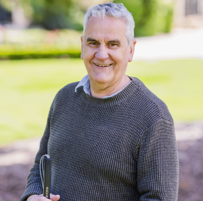 An older man with grey hair, wearing a grey knit jumper, stands outdoors on a sunny day, smiling and holding a golf club on a lush green course. The background is blurred grass and trees.