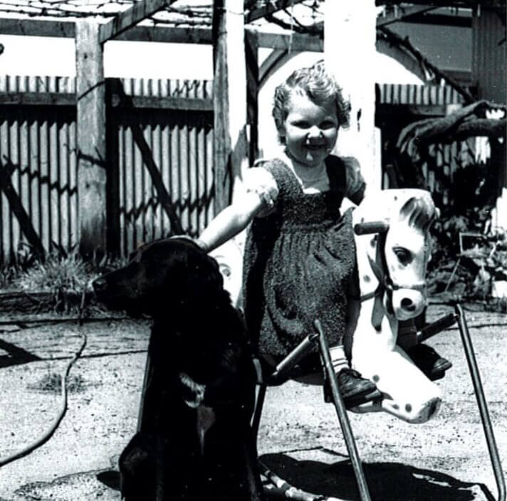A young girl in a dress sits on a spring rocking horse outdoors, smiling and holding a black dog beside her. A wooden fence and trees are visible in the background.