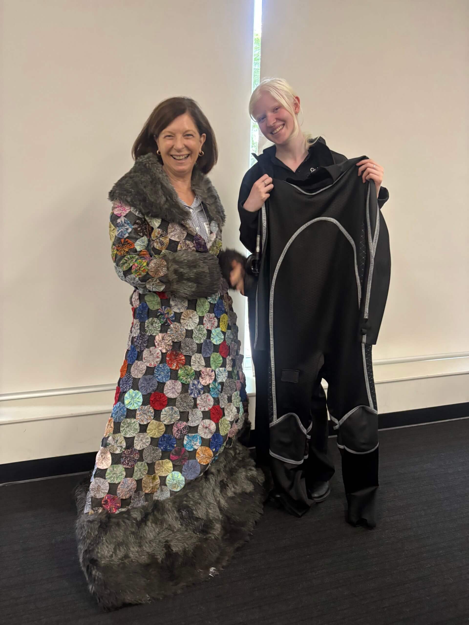 Two women stand indoors smiling. One wears a long, colourful patchwork coat with fake fur trim, while the other holds a large black garment with grey trim. A window is partially visible behind them.