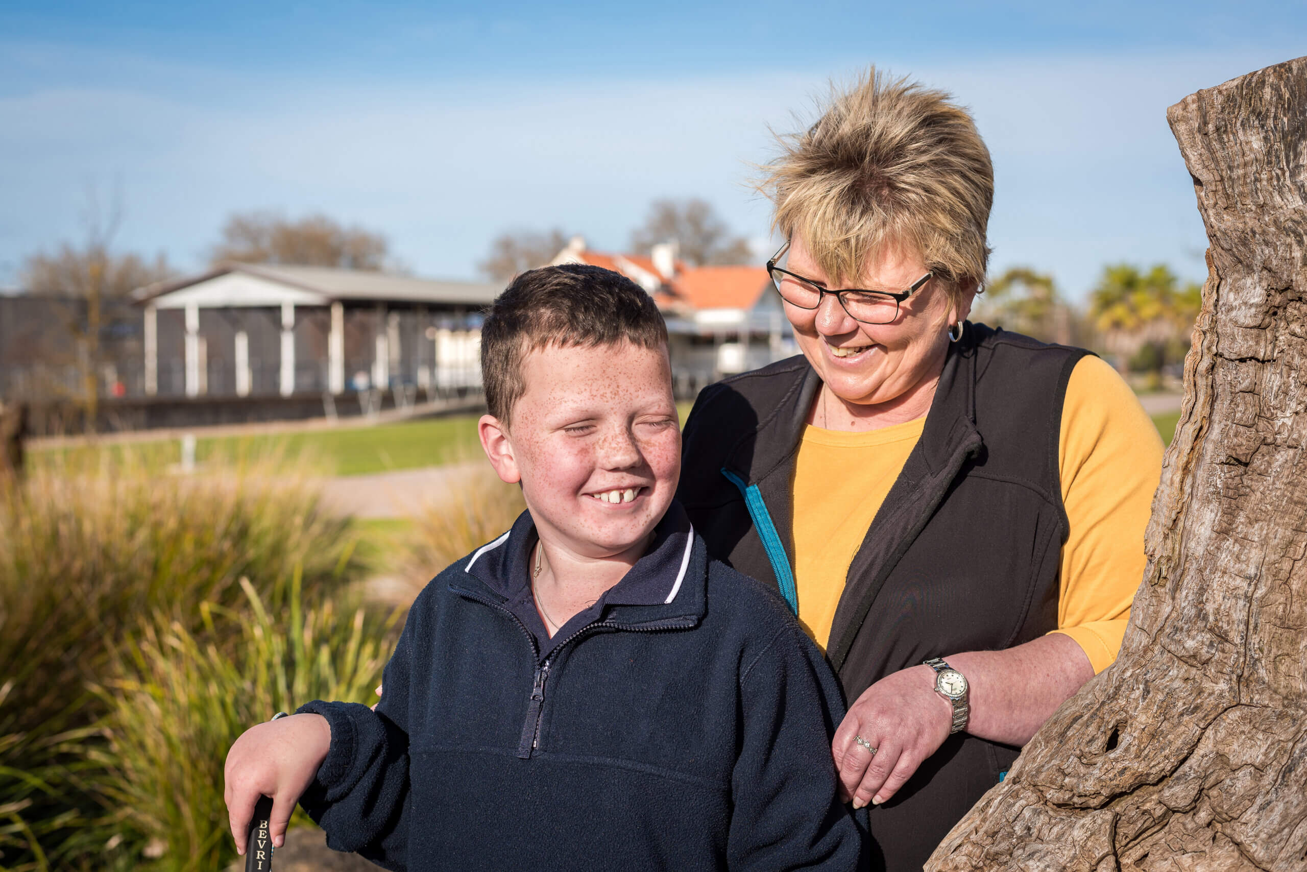 A smiling boy stands outdoors next to a tree, holding a stick, while an older woman in glasses and a yellow shirt stands behind him, smiling warmly at him. There are buildings and greenery in the background.