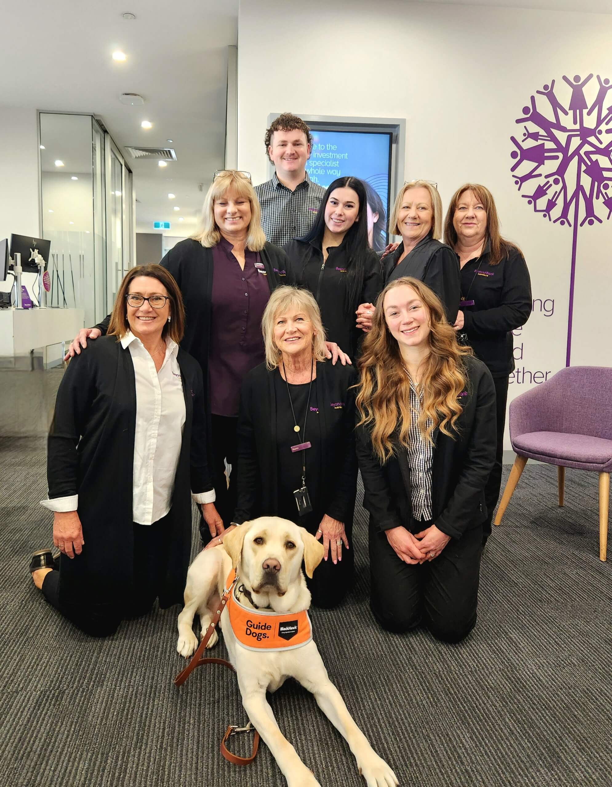 A group of eight smiling adults, mostly women, pose indoors with a guide dog lying at the front. The people wear business attire and stand in an office with modern décor and a purple tree mural on the wall.