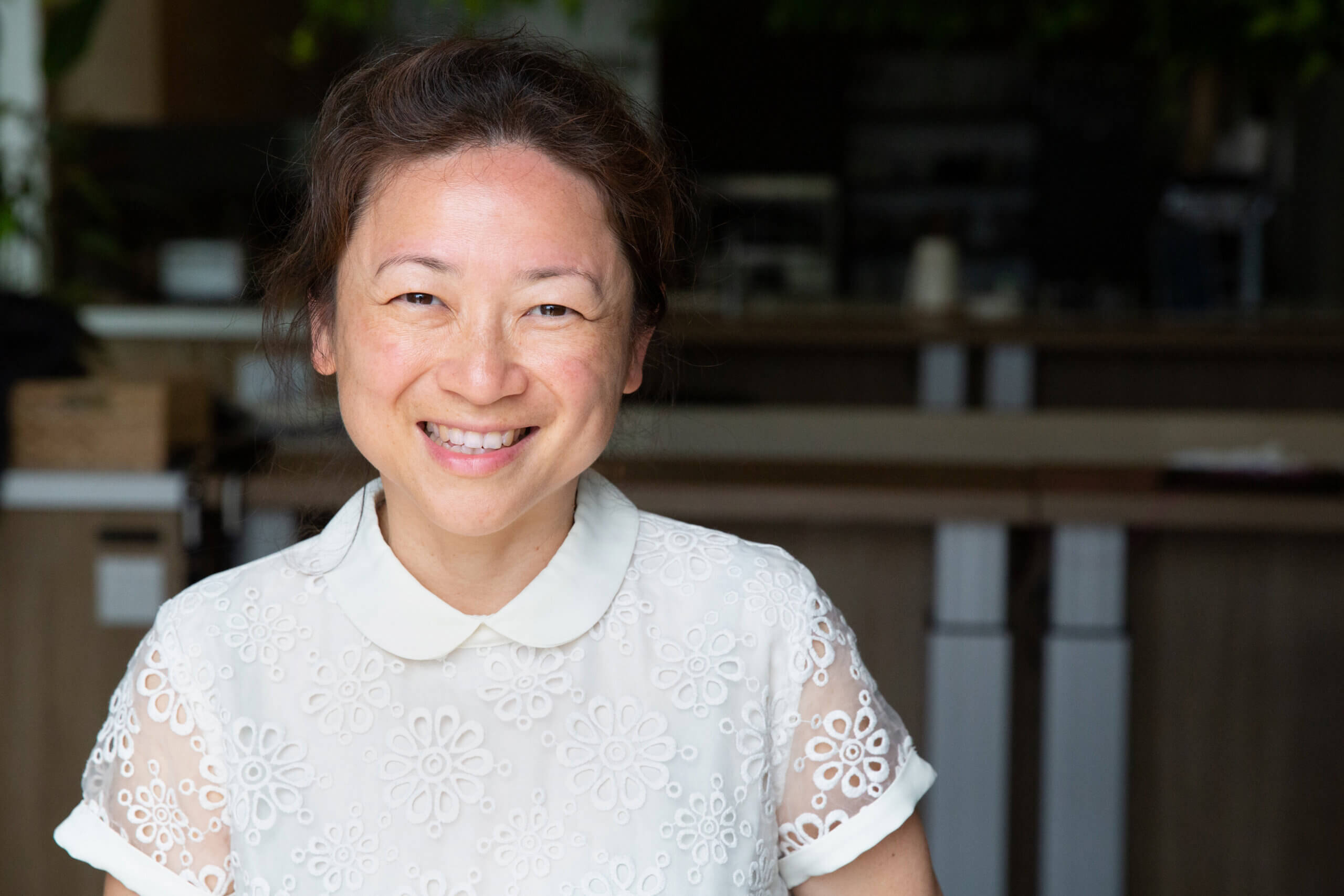 A woman with dark hair tied back, wearing a white lace blouse with a collar, smiles warmly at the camera whilst sitting indoors in a bright, modern setting, exuding elegance.