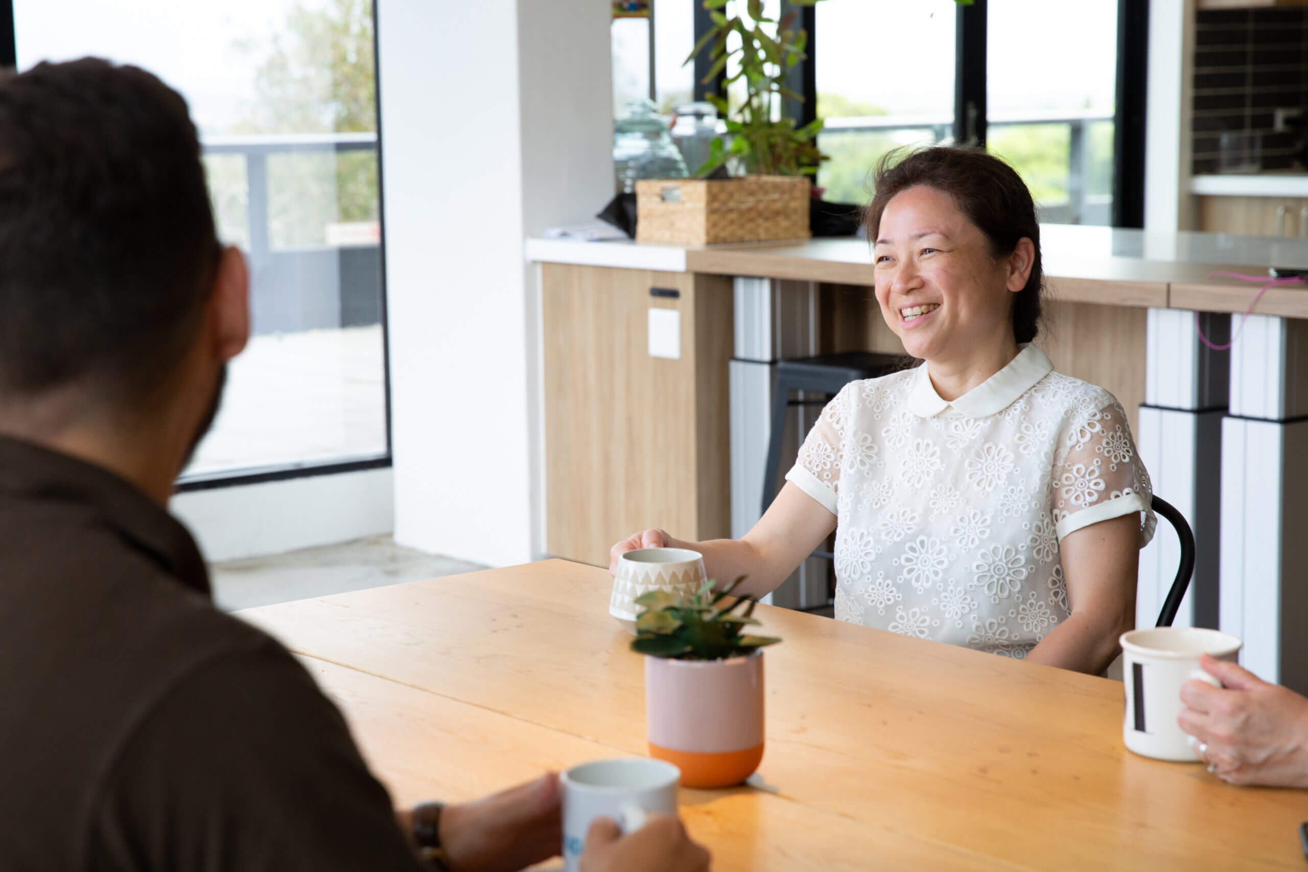 A woman smiles whilst sitting at a wooden table with two others, holding a mug. They enjoy a casual conversation in a bright, modern office space with large windows and a potted plant on the table.