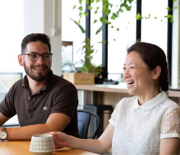 Two people sit at a table, smiling and talking. The woman in the foreground holds a mug, whilst the man in glasses listens. Sunlight streams through large windows, highlighting the greenery from potted plants nearby.