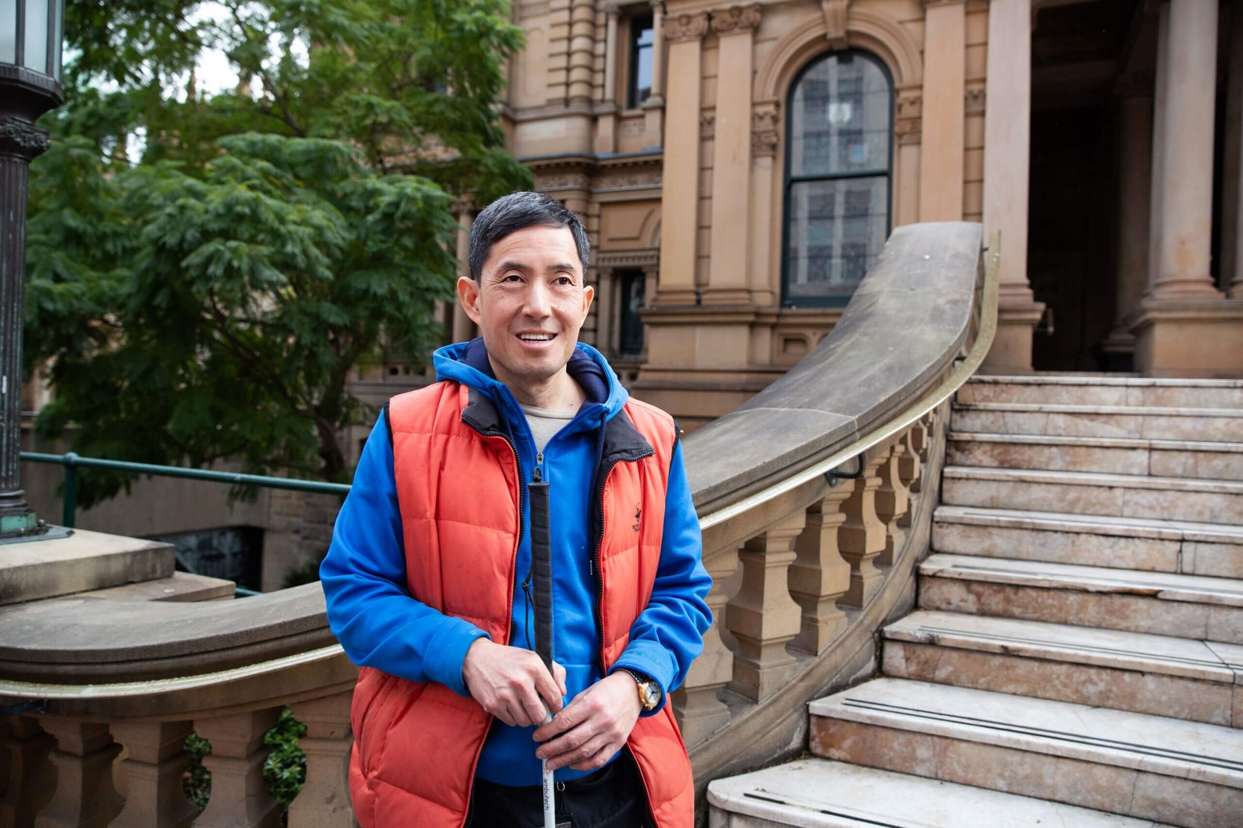 A man wearing a blue hoodie and orange vest stands smiling in front of a curved stone staircase and a historic building with large windows and ornate architecture. Trees are visible in the background.