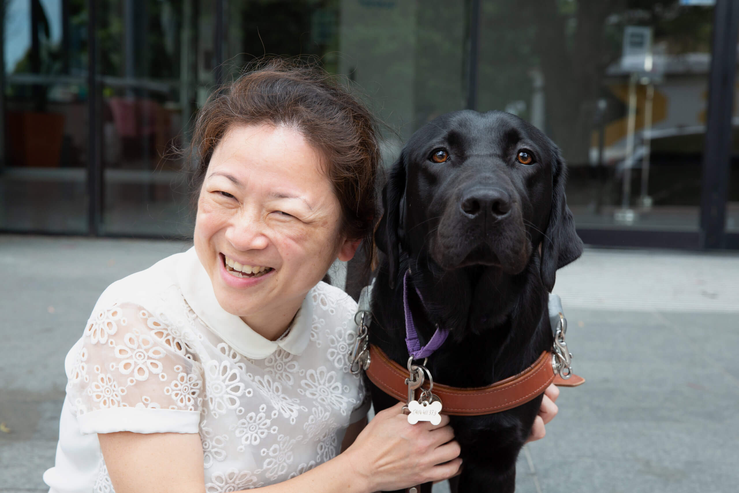 A smiling woman crouches next to a black guide dog wearing a brown harness and purple collar, outdoors in front of a glass building.