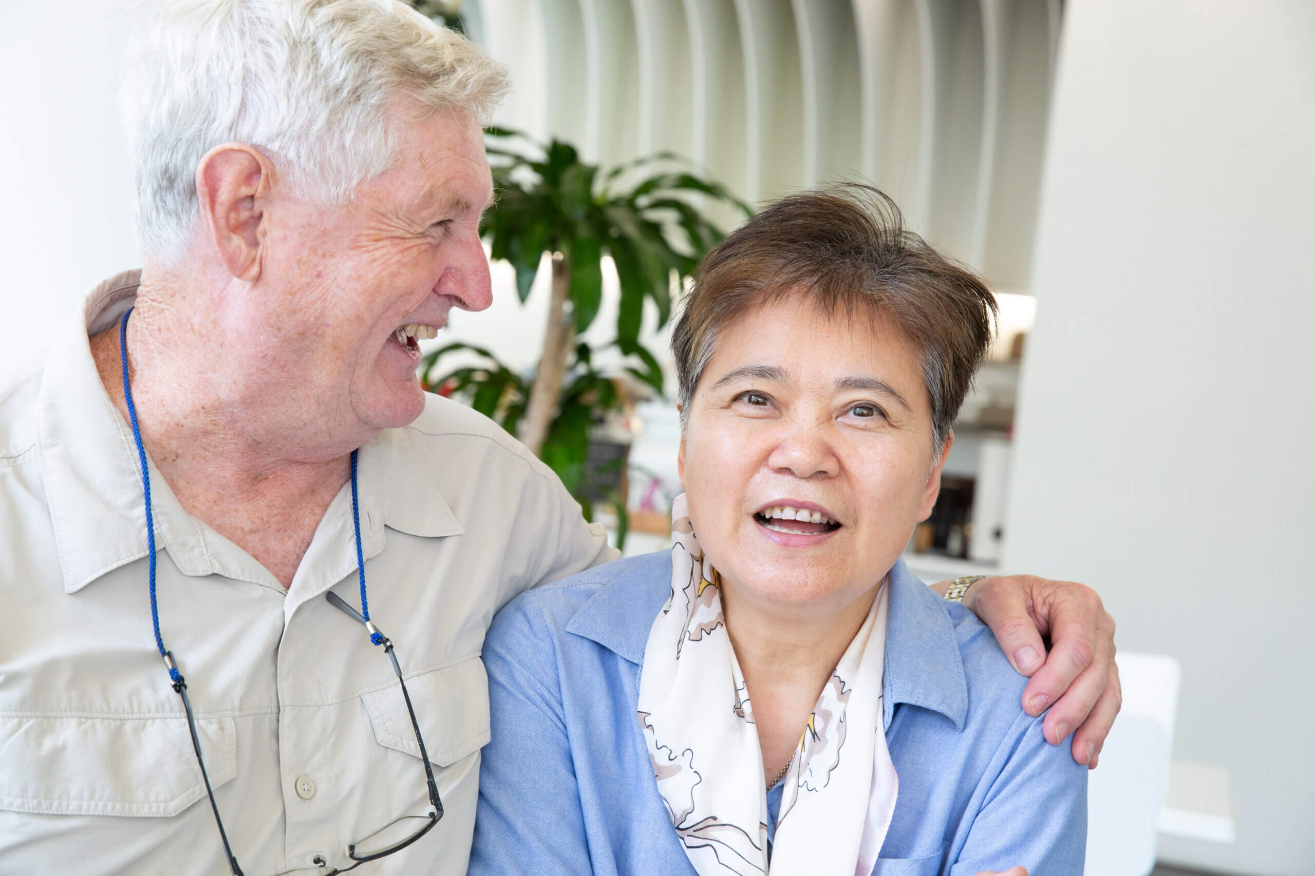 An older man smiles at an older woman while gently putting his arm round her shoulders. The woman, wearing a light blue shirt and scarf, looks towards the camera and smiles slightly. They are indoors with a plant in the background.