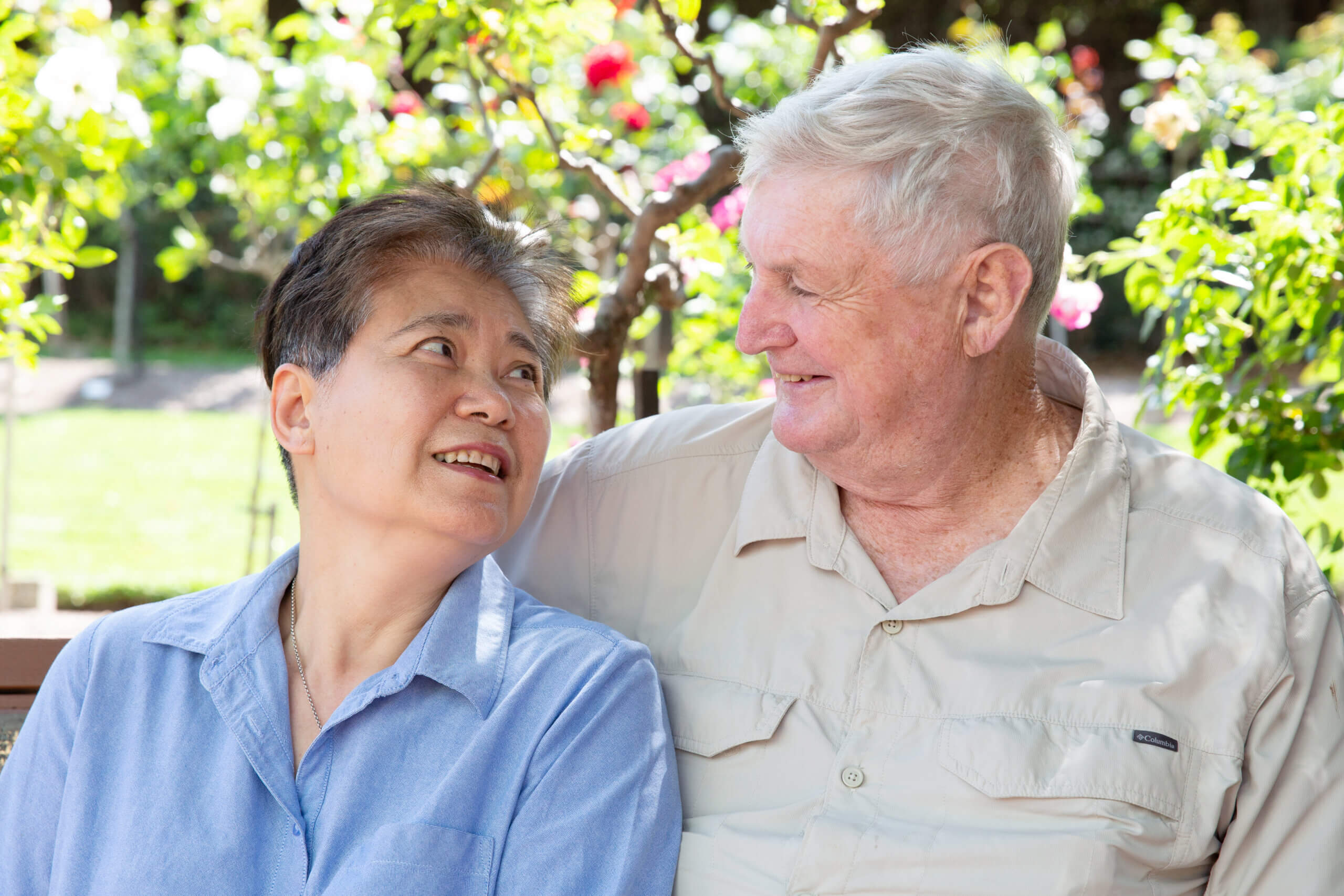 An older couple sit closely together on a bench outdoors, smiling warmly at each other. Greenery and colourful flowers fill the background, creating a peaceful, sunny setting.