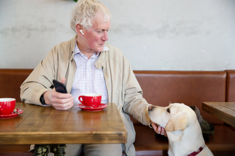 An older man sits at a café table with a red coffee cup, holding a mobile and gently stroking a yellow Labrador sitting beside him. The man looks at the dog with a fond expression.
