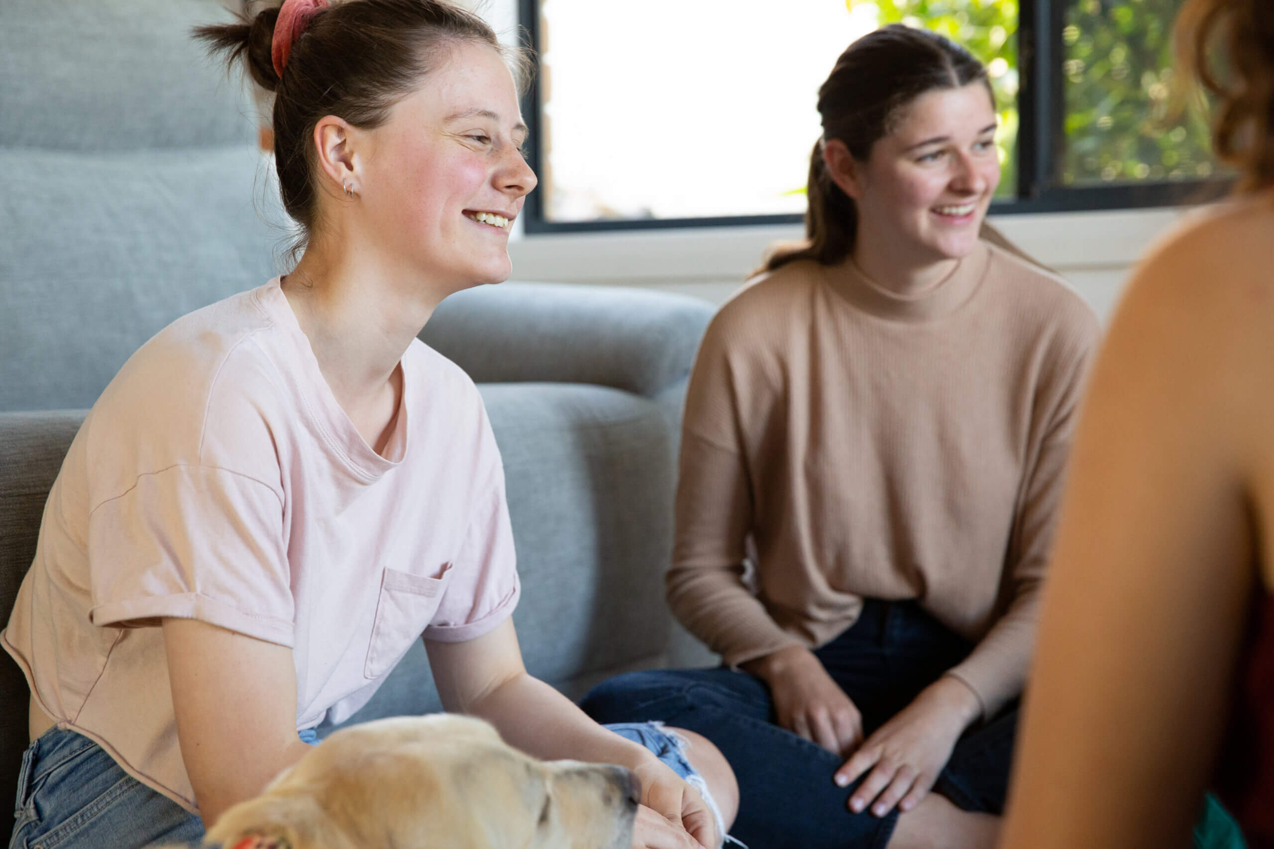 Two young women sit on the floor indoors, smiling and talking beside a dog. One wears a light pink shirt, the other a beige top and jeans. Natural light streams through a window, creating a warm, inviting atmosphere.