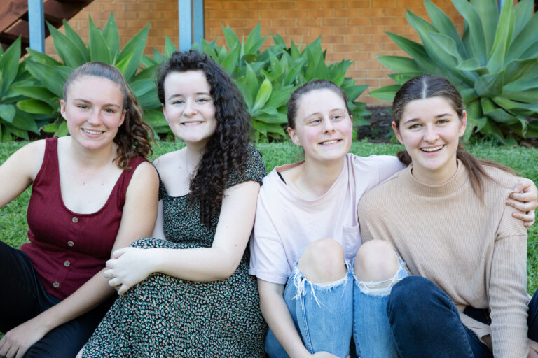 Four young women sit close together on grass, smiling at the camera. Behind them are green plants and a brick wall. They wear casual clothes in shades of red, green, pink, and beige.