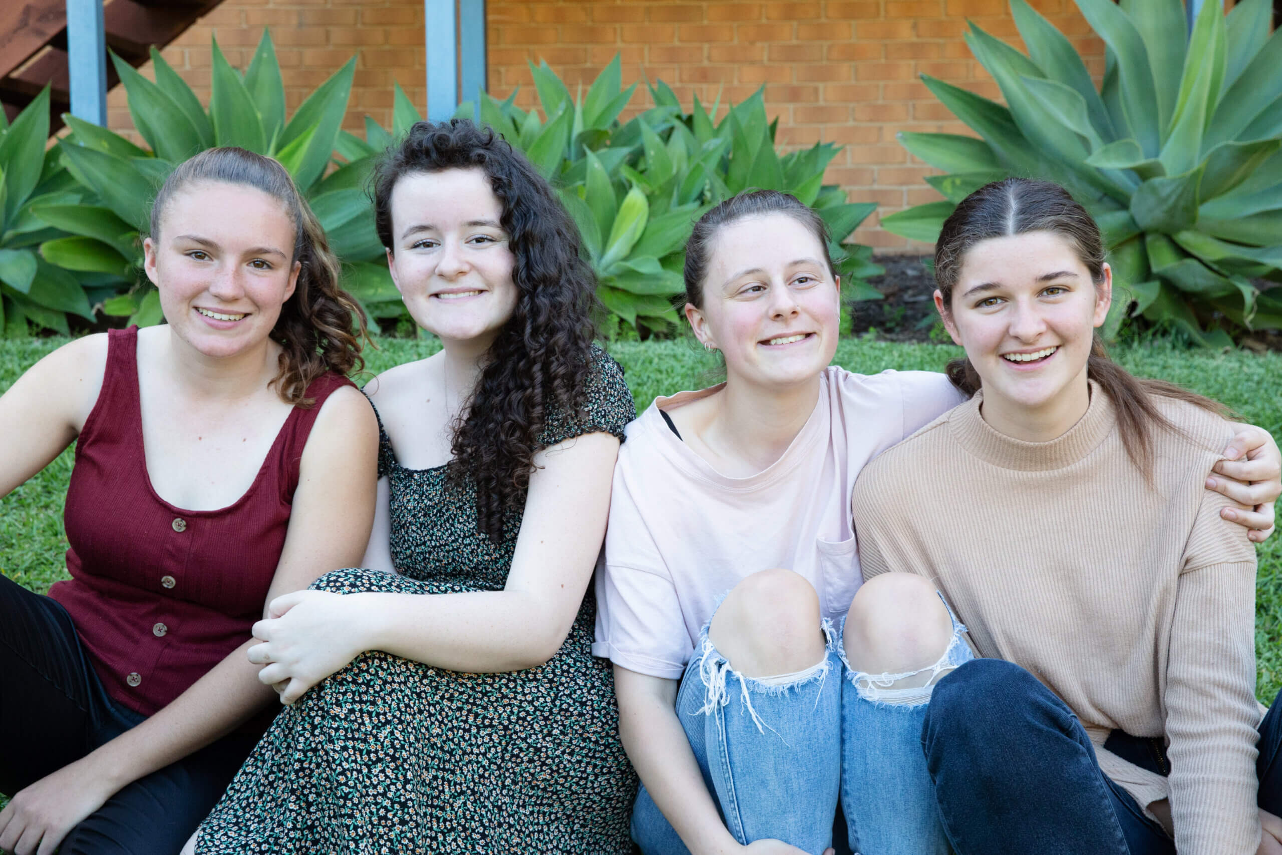 Four young women sit closely together on grass, smiling at the camera. Behind them are large green plants and a brick wall. They wear casual clothes, including a red sleeveless top, floral dress, white T-shirt, and beige jumper.