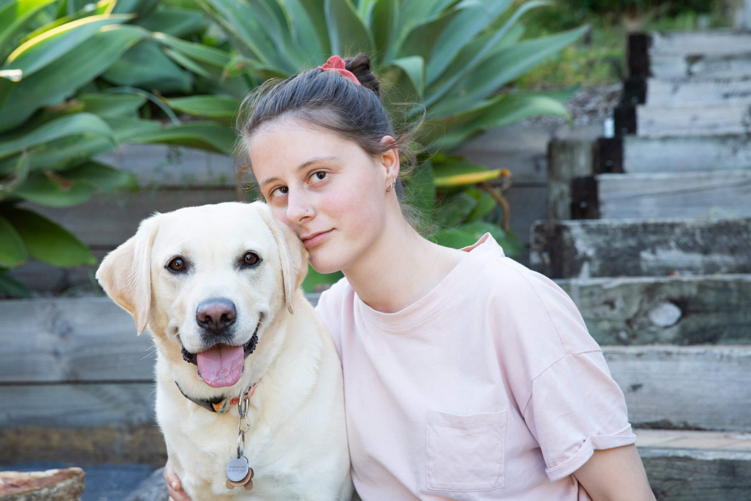 A young woman with brown hair in a ponytail sits next to a smiling yellow Labrador retriever. They are outdoors on wooden steps, surrounded by green leafy plants. The woman wears a light pink T-shirt and enjoys time with her loyal companion.