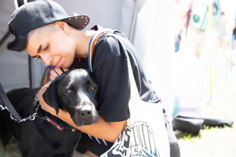 A person wearing a backwards cap and a rainbow-strap bag hugs a black dog closely, both appearing calm and affectionate. The scene is outdoors and brightly lit.