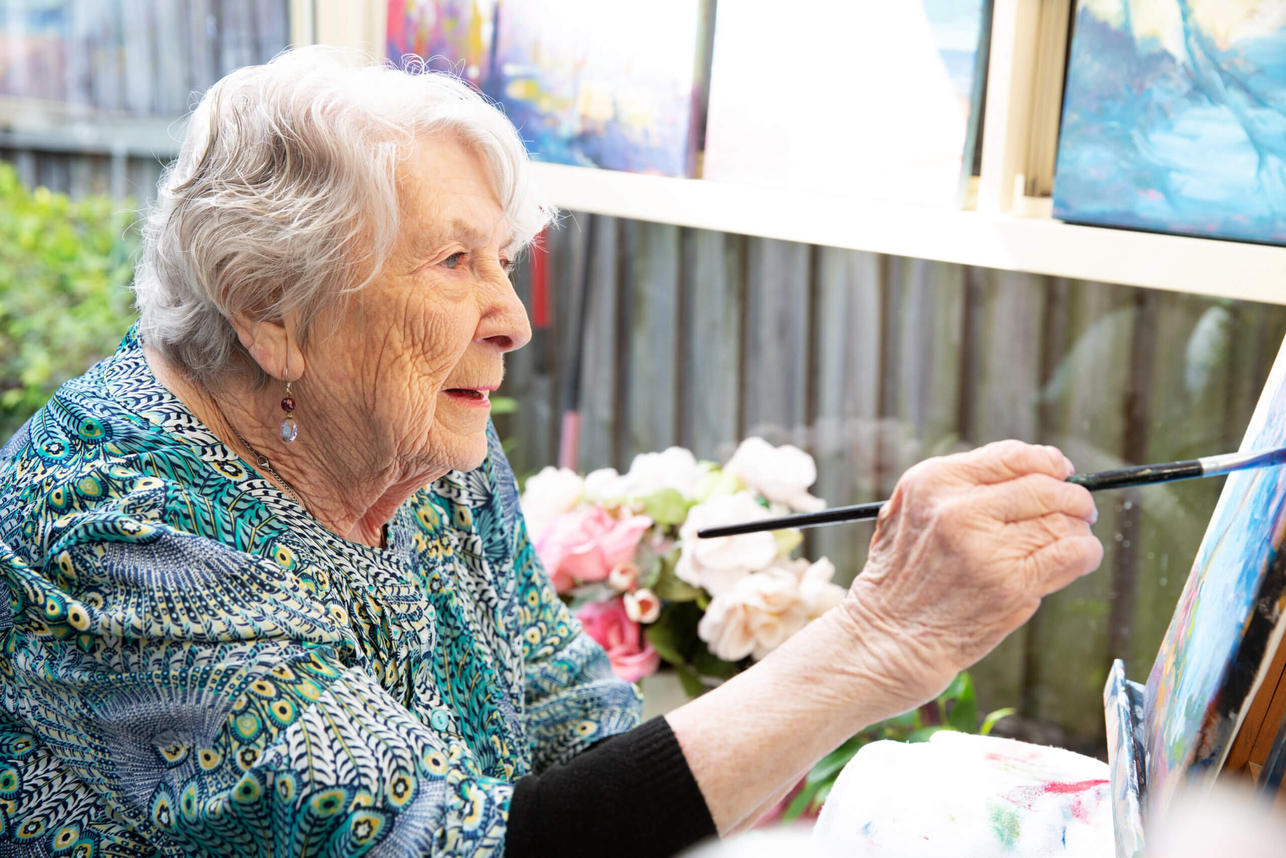 An older woman with grey hair paints on a canvas, smiling and focused. She wears a blue patterned top and is surrounded by flowers and colourful paintings in a bright room.