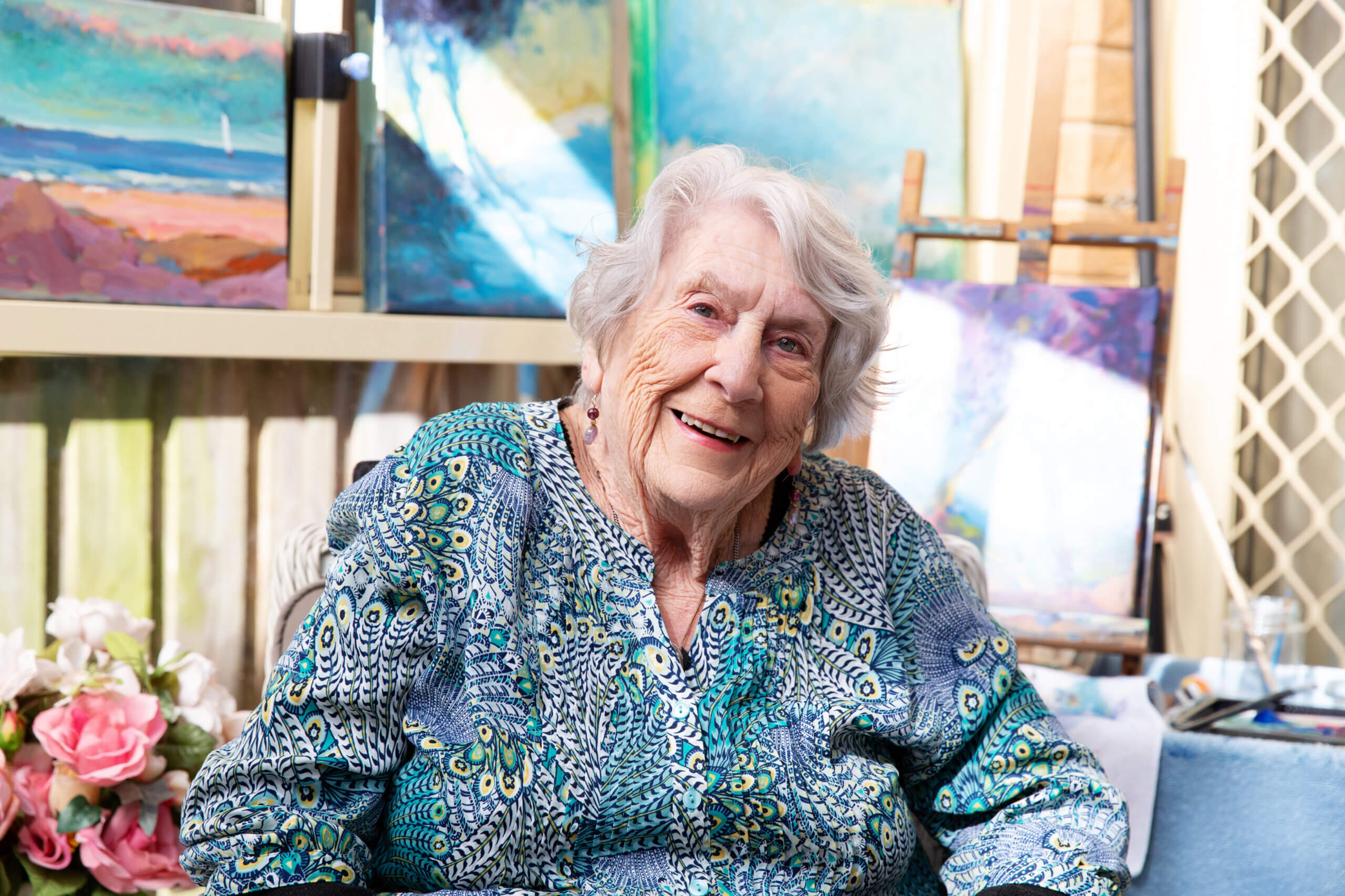 An elderly woman with white hair, wearing a blue patterned blouse, smiles whilst sitting indoors. Behind her are colourful paintings, an easel, and a bunch of pink flowers, creating a creative and artistic atmosphere.