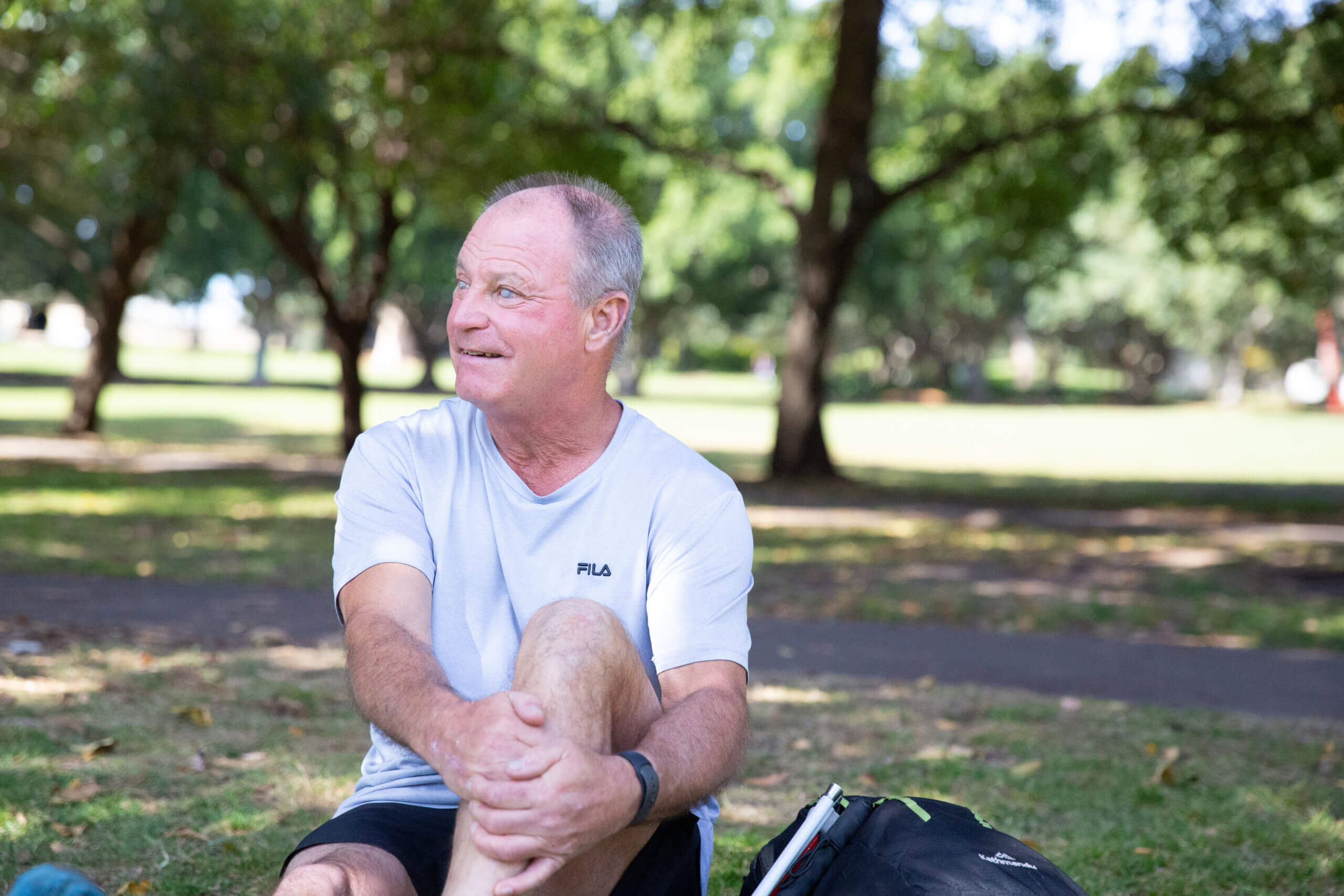 An older man wearing a white FILA T-shirt sits on grass in a park, smiling and looking to the side. Trees and sunlight create a relaxed atmosphere, while his black bag rests beside him.