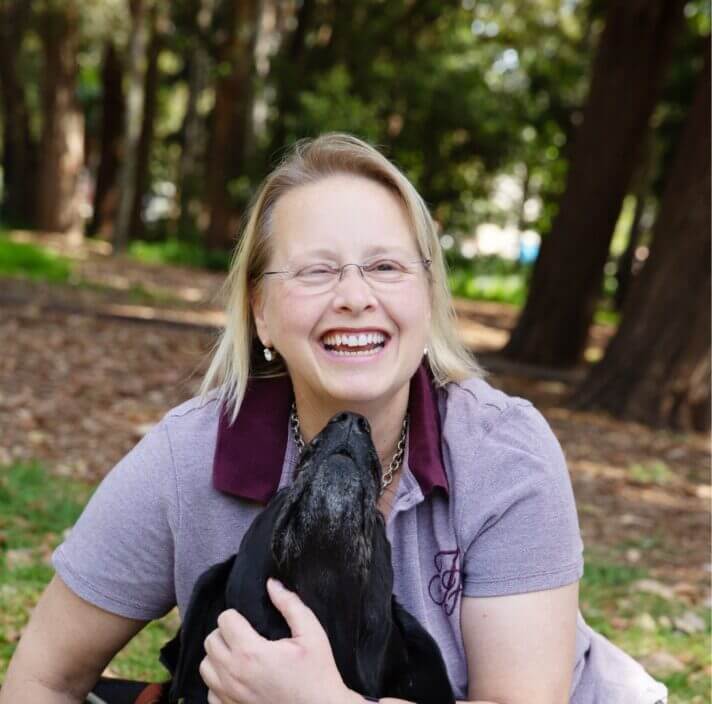 A woman with glasses and blonde hair smiles joyfully whilst sitting outdoors in a wooded area, as her black dog affectionately nuzzles her neck.