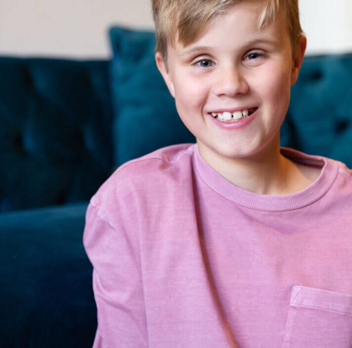Smiling blonde-haired child in a pink shirt sits happily in front of a dark blue settee.