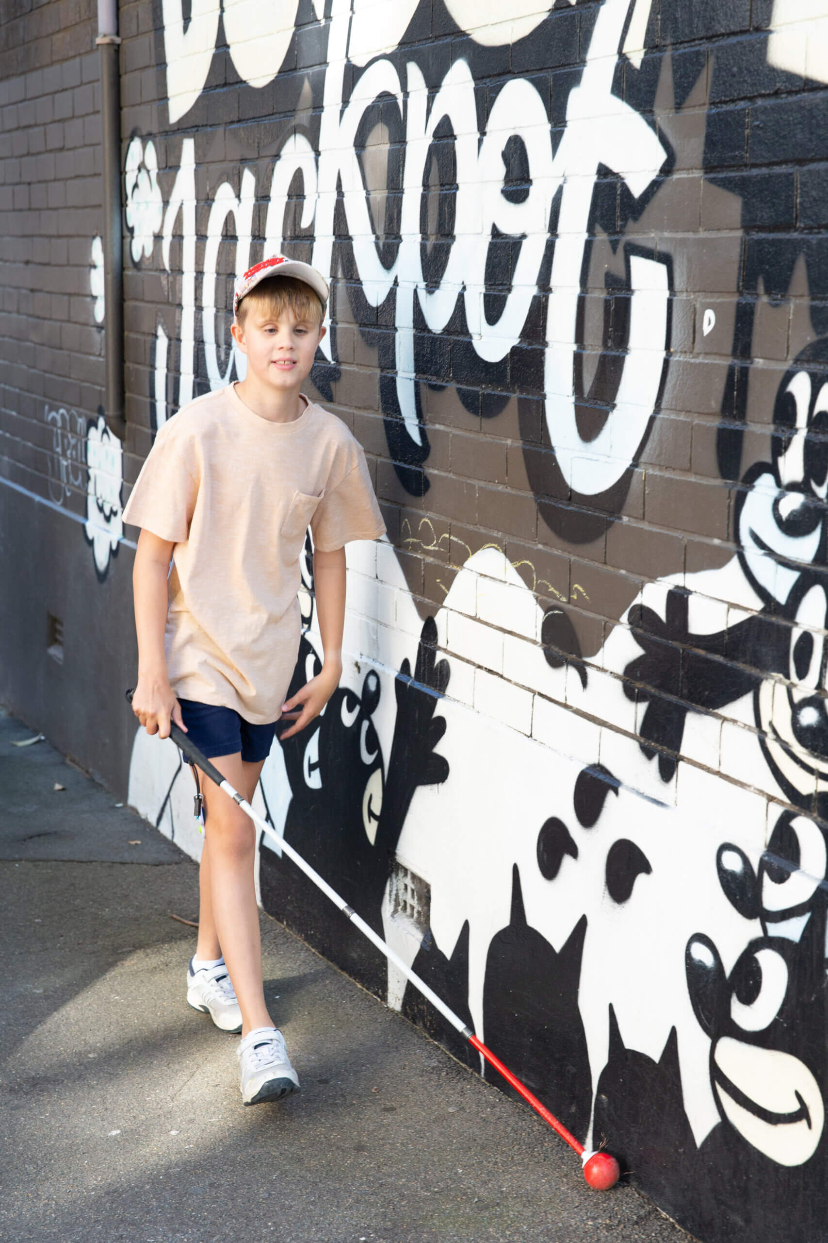 A boy wearing a cap and holding a white cane walks alongside a mural-covered brick wall with bold black and white graffiti art.