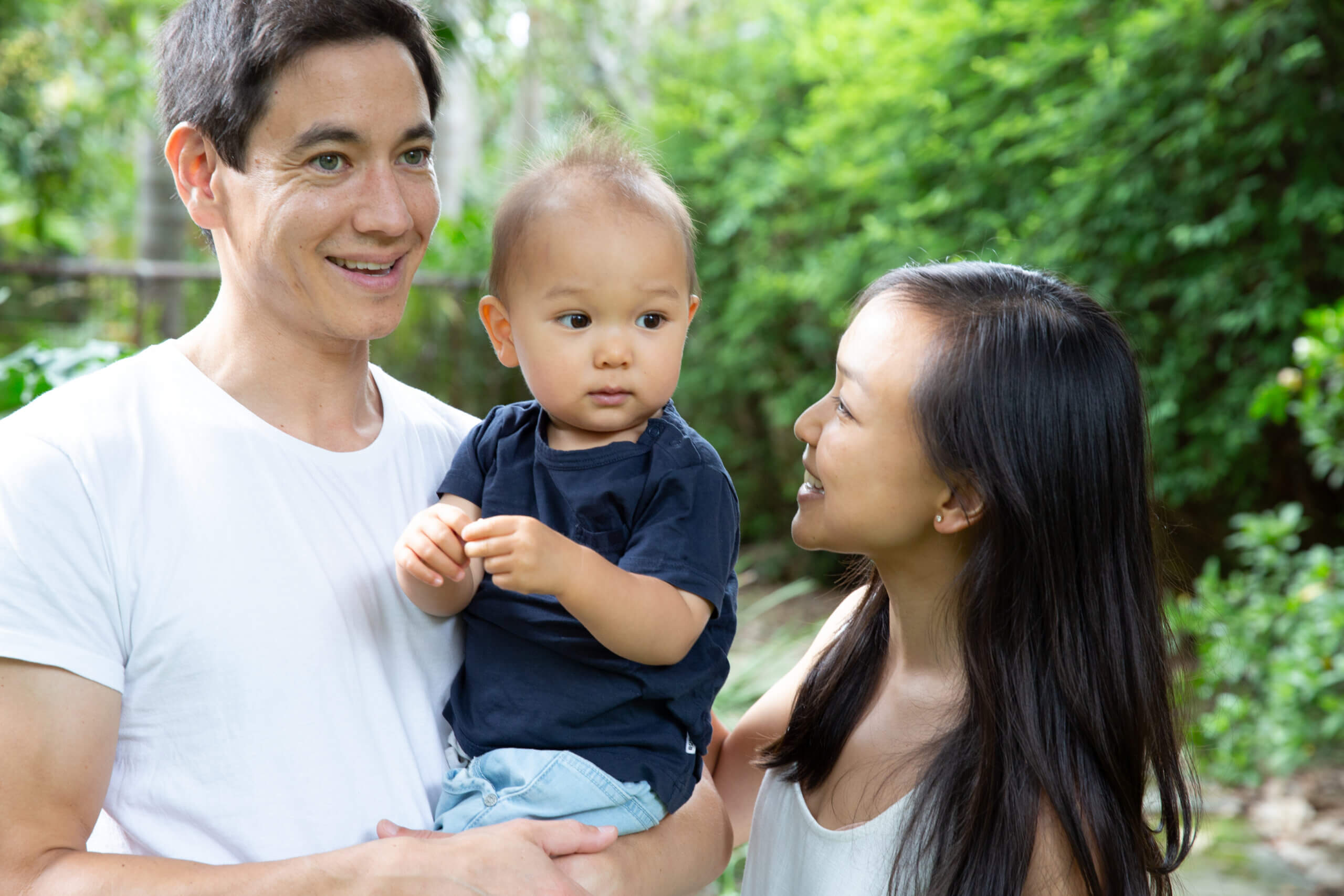 A man holds a baby while standing next to a woman who smiles at them. They are outdoors with green foliage in the background, and all three are casually dressed.