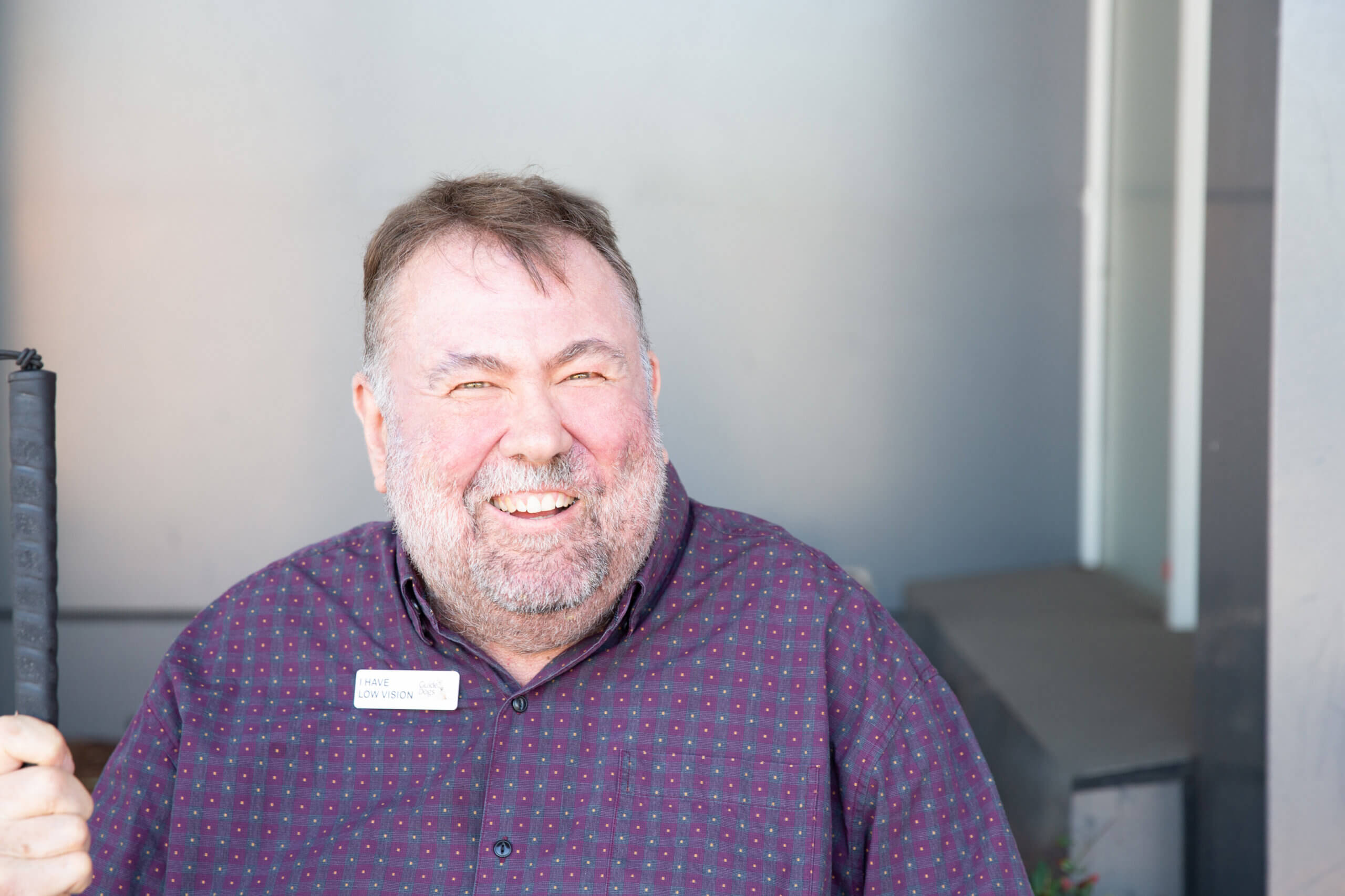 A smiling man with a beard wears a maroon patterned shirt and a name badge, standing in front of a grey background. He is holding a black walking stick in his left hand.