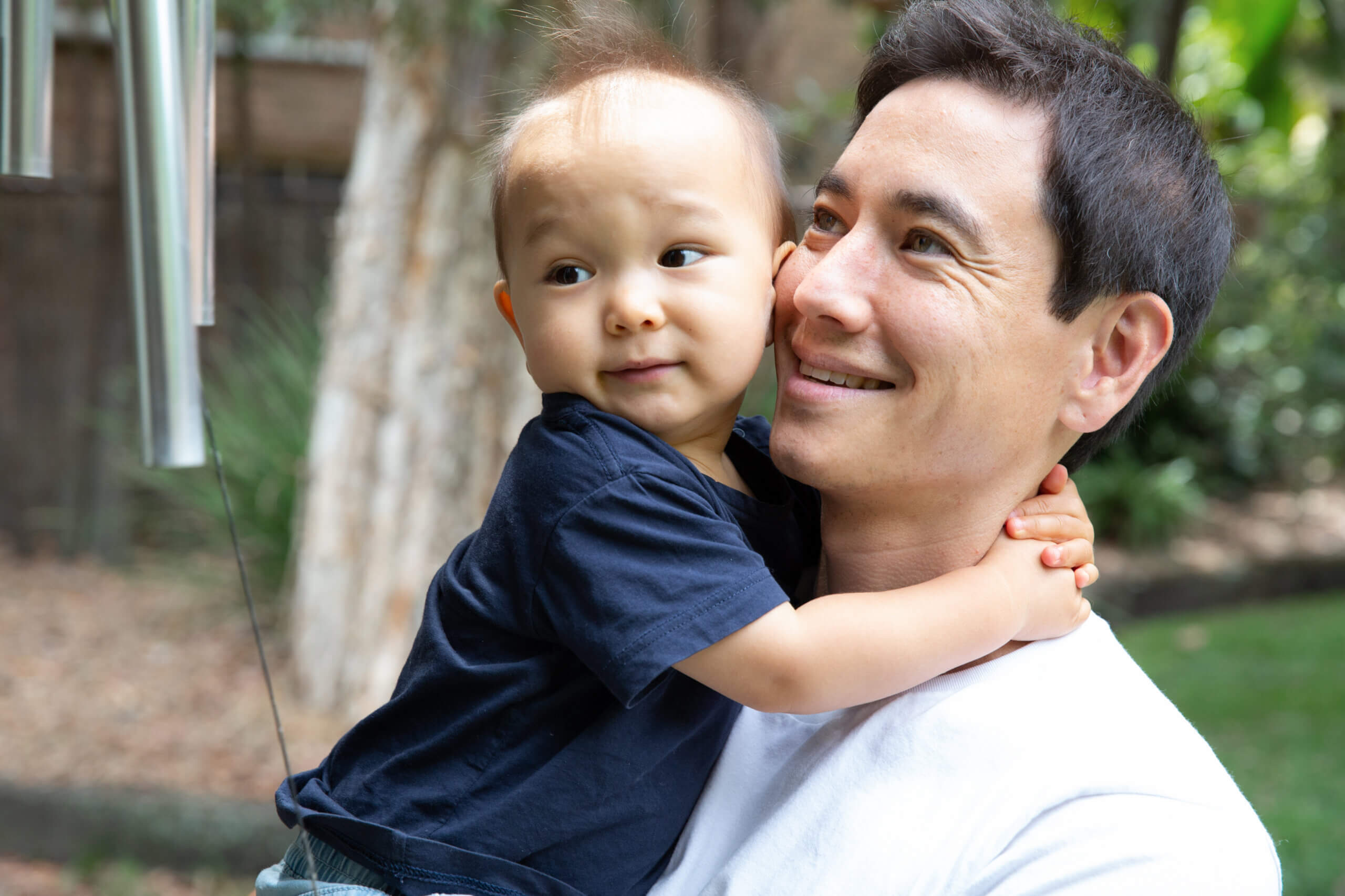 A man smiles whilst holding a young child in his arms outdoors. The child looks off to the side, one arm around the man's neck. Trees and lush greenery create a serene background.