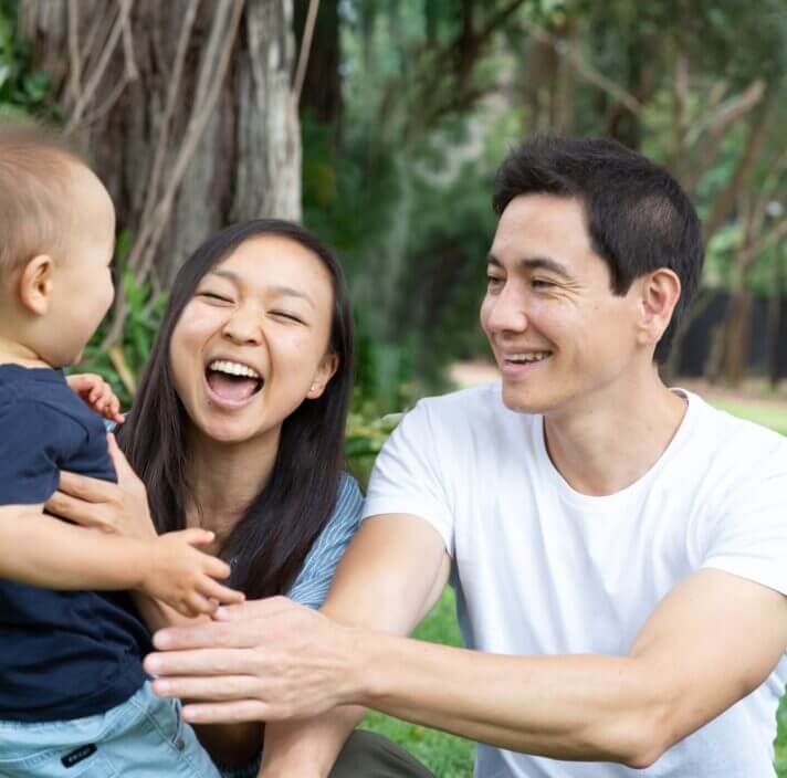 A smiling couple sits outdoors on grass, reaching out to their young child who stands in front of them. Surrounded by trees and greenery, the family enjoys quality time together, looking happy and engaged.