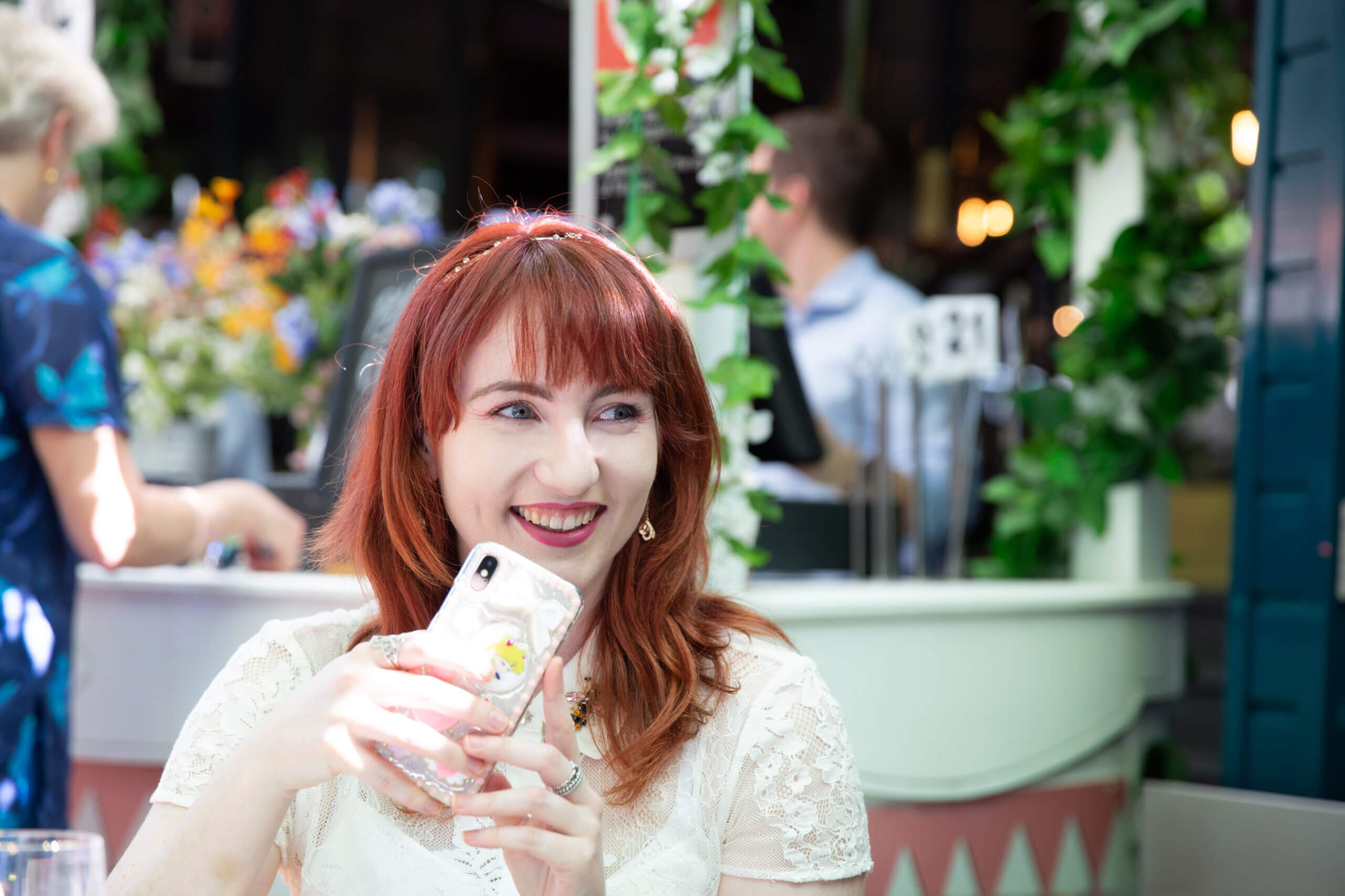 A woman with red hair in a white dress sits at a table, smiling and holding a mobile. She is outdoors with lush greenery and people in the background, enjoying a bright, sunny day.