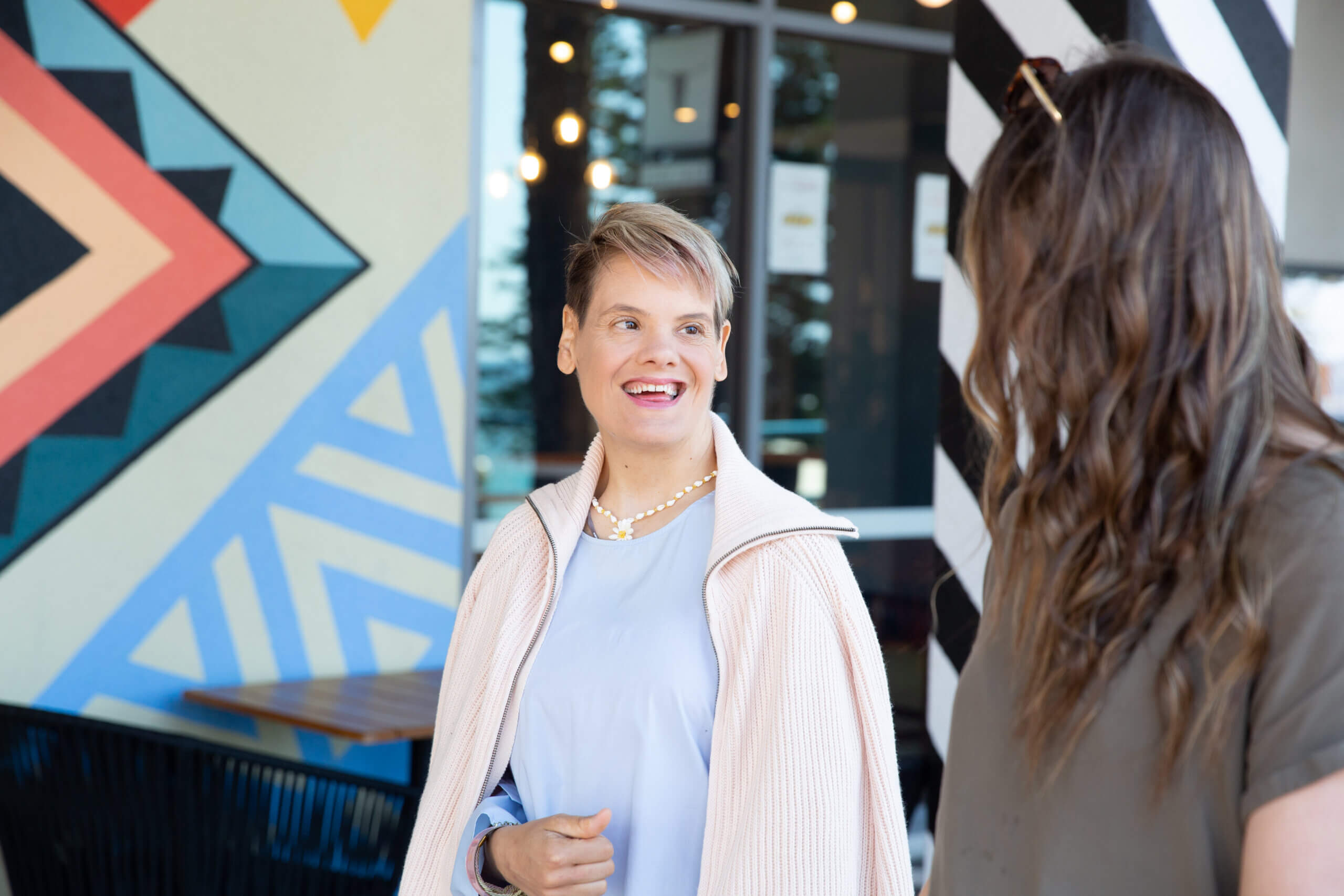 Two women stand and converse outside a building with colourful geometric wall art. One woman smiles, wearing a light top and a necklace, whilst the other has long wavy hair and sunglasses on her head.