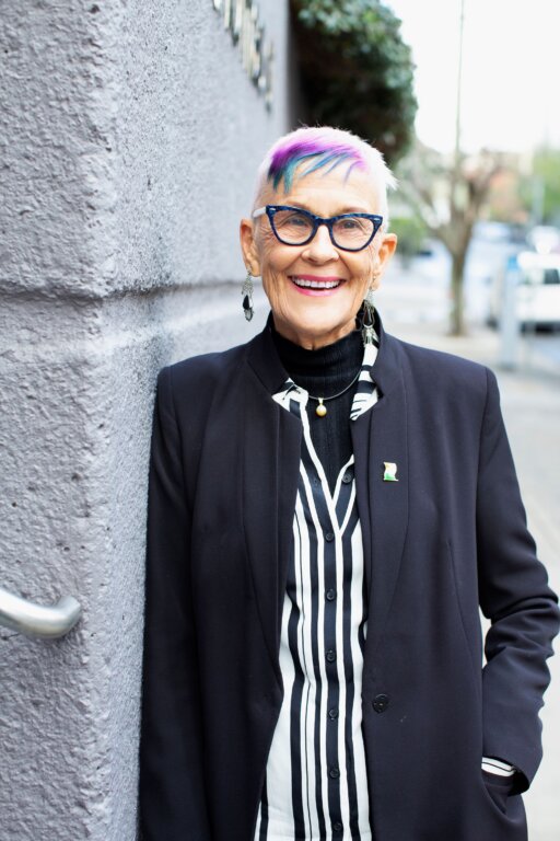 An older person with short white hair dyed purple and blue at the front, wearing blue glasses, a dark blazer, and a black-and-white striped shirt, smiles whilst standing against a grey wall outdoors.