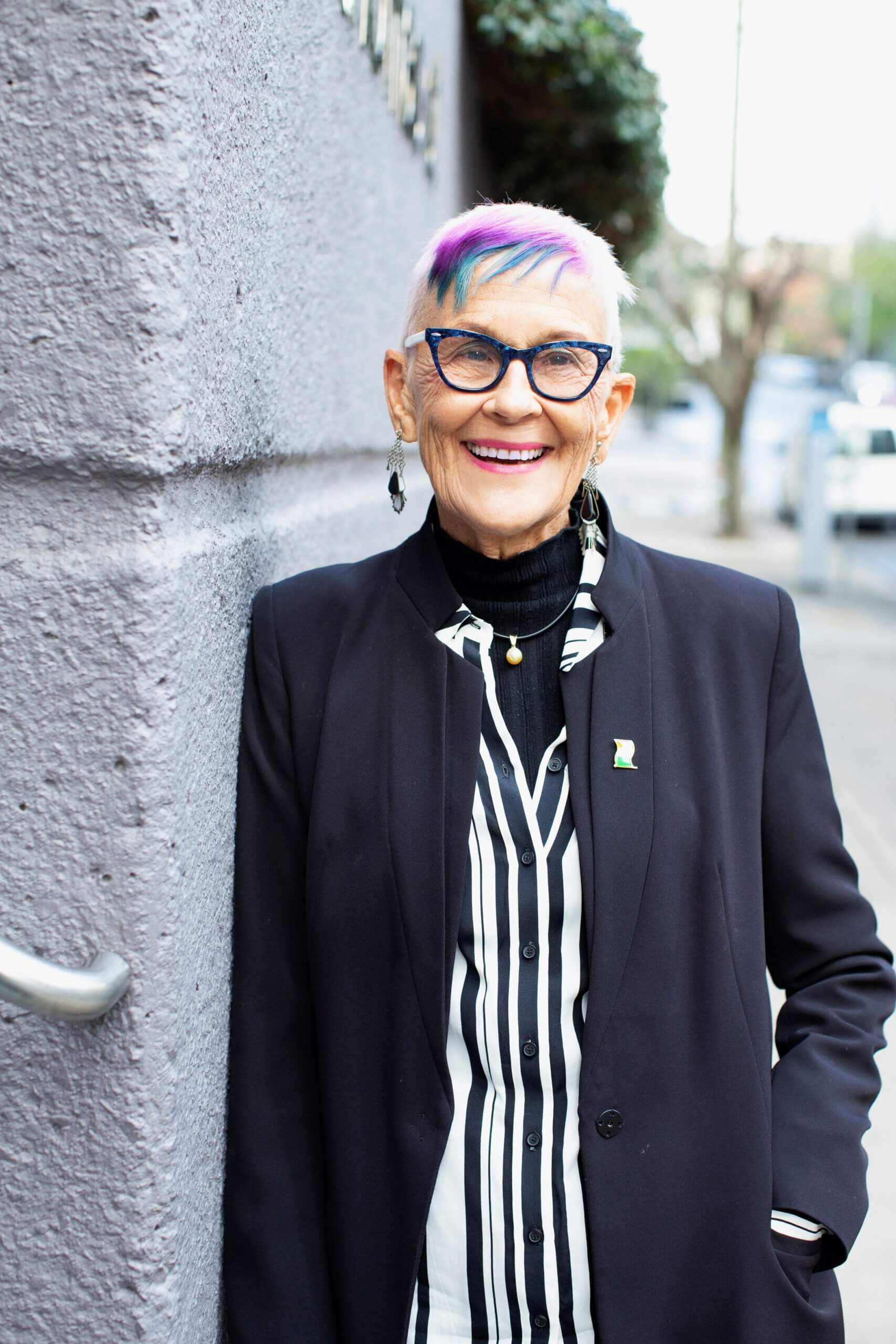 An older person with short white hair dyed purple and blue at the front, wearing blue glasses, a dark blazer, and a black-and-white striped shirt, smiles whilst standing against a grey wall outdoors.