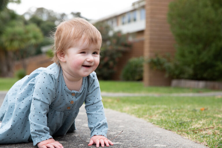 A smiling baby in a light blue polka dot outfit crawls on a concrete path outdoors, with lush grass, bushes, and a building in the background.