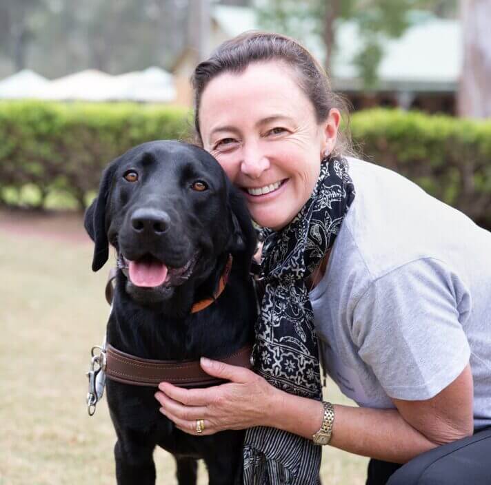 A smiling woman kneels beside a joyful black Labrador in a harness, outdoors on a grassy area surrounded by lush green bushes and trees.