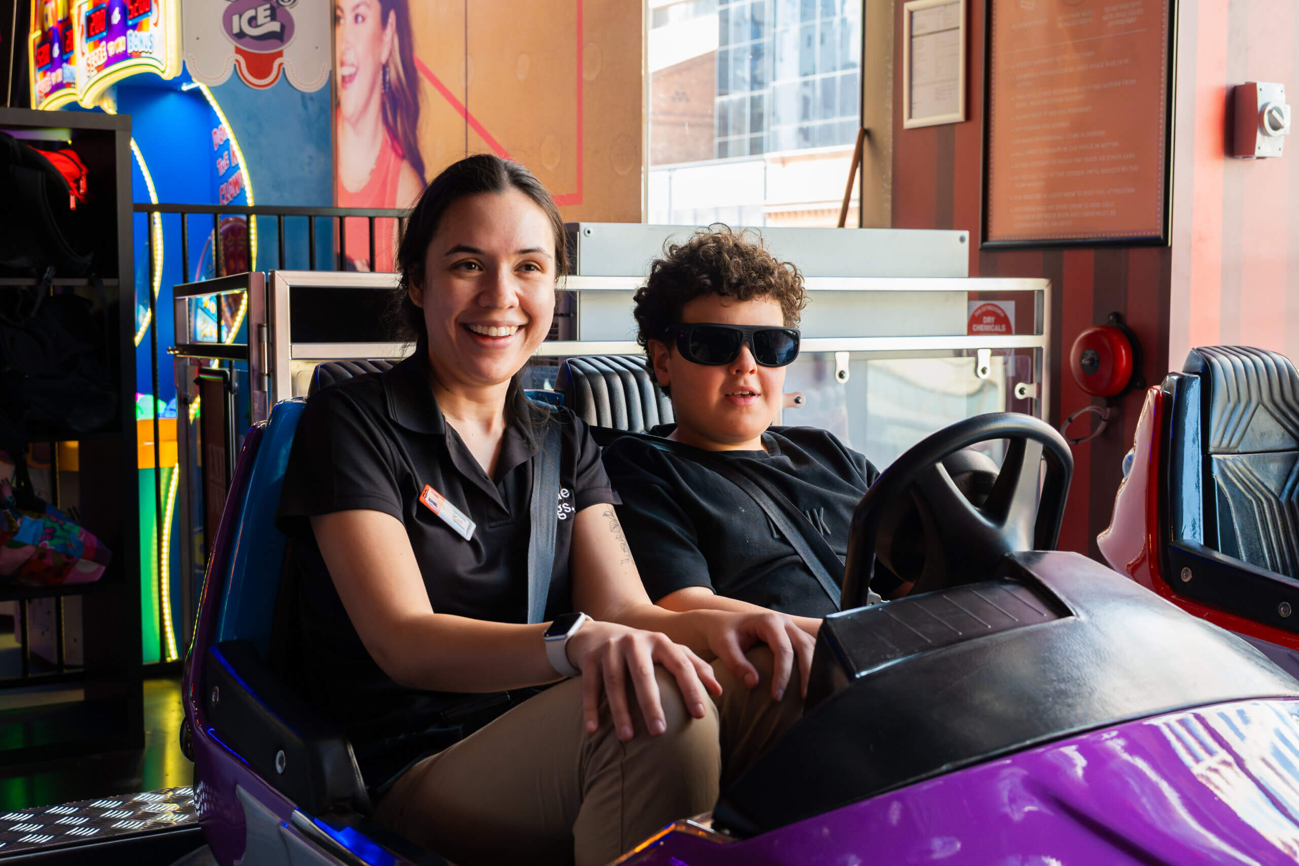 A woman and a boy wearing sunglasses sit in a purple dodgem car, smiling. Bright neon lights and vibrant arcade games fill the background.